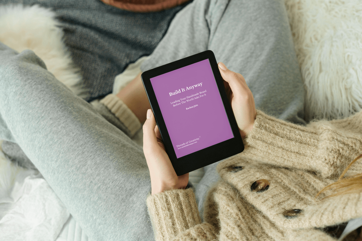 A creative founder in cozy clothes sits cross-legged, holding an e-reader that displays “Build It Anyway: Leading Your Handmade Brand” from Mirabilia Boutique—encouraging launching ideas and leading without waiting for approval.