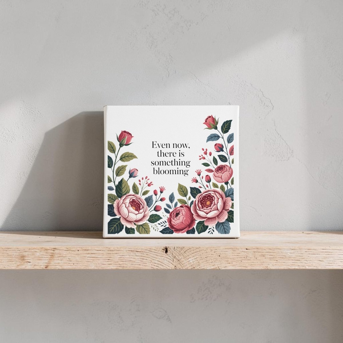 The Even Now There Is Something Blooming Canvas Wall Art by Mirabilia Boutique features pink and red florals with inspiring text—ideal as an encouragement gift and lovely displayed on a light shelf against a soft, textured wall.