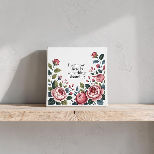 The Even Now There Is Something Blooming Canvas Wall Art by Mirabilia Boutique features pink and red florals with inspiring text—ideal as an encouragement gift and lovely displayed on a light shelf against a soft, textured wall.