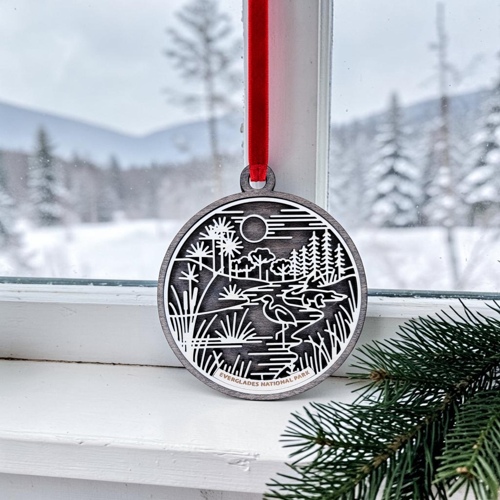 A Mirabilia Boutique Everglades National Park Line Art Christmas Ornament, featuring an engraved silver nature scene, hangs by a red ribbon in front of a snowy window while evergreen branches accent the corner.