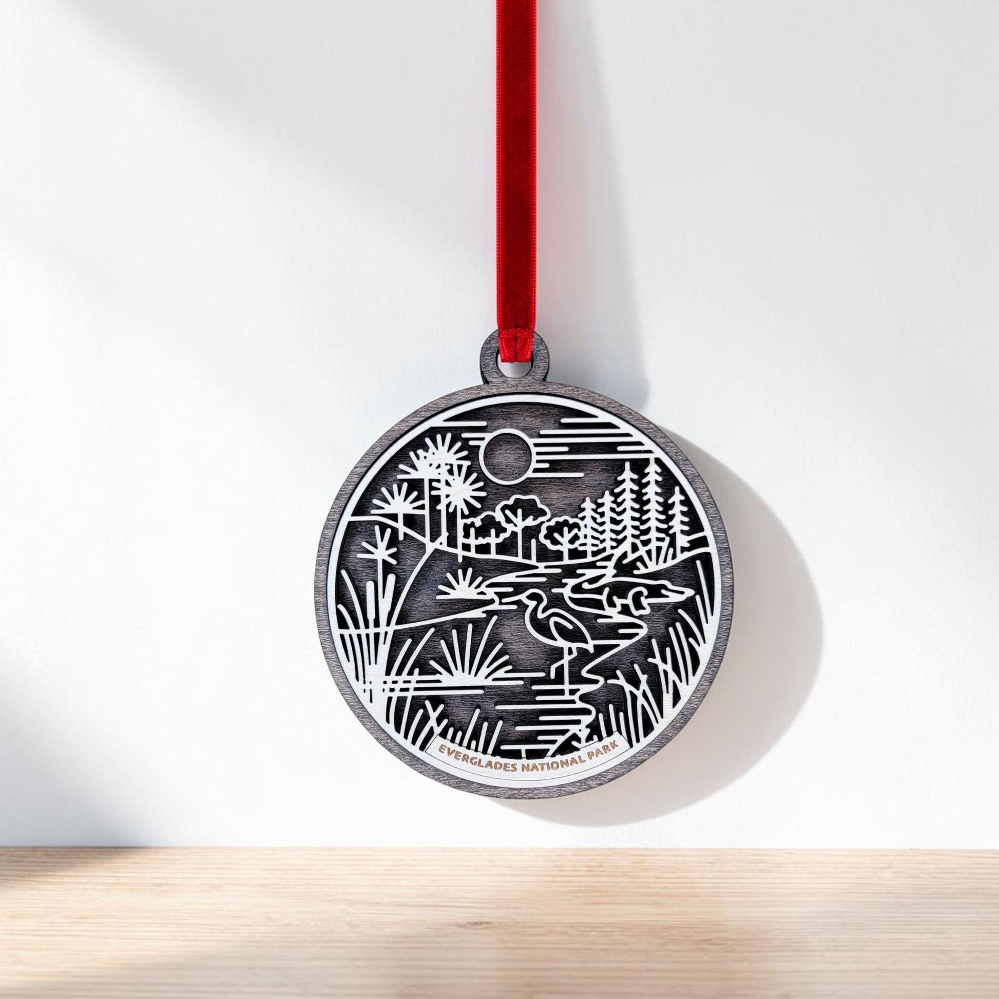 A round silver Everglades National Park Line Art Christmas Ornament by Mirabilia Boutique, showcasing a nature scene with trees, water, and a moon, hangs from a red ribbon against a white wall above a light wooden surface.
