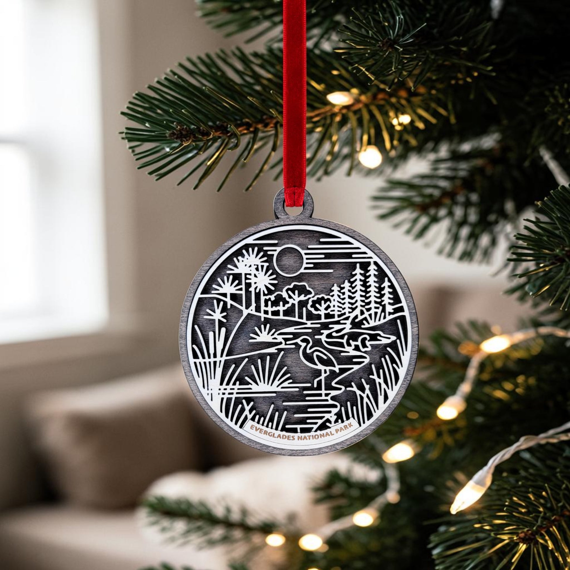 A Mirabilia Boutique Everglades National Park Line Art Christmas Ornament, handcrafted from wood and featuring an engraved scenic park motif, hangs from a red ribbon on a lit Christmas tree; a blurred couch and window are visible in the background.