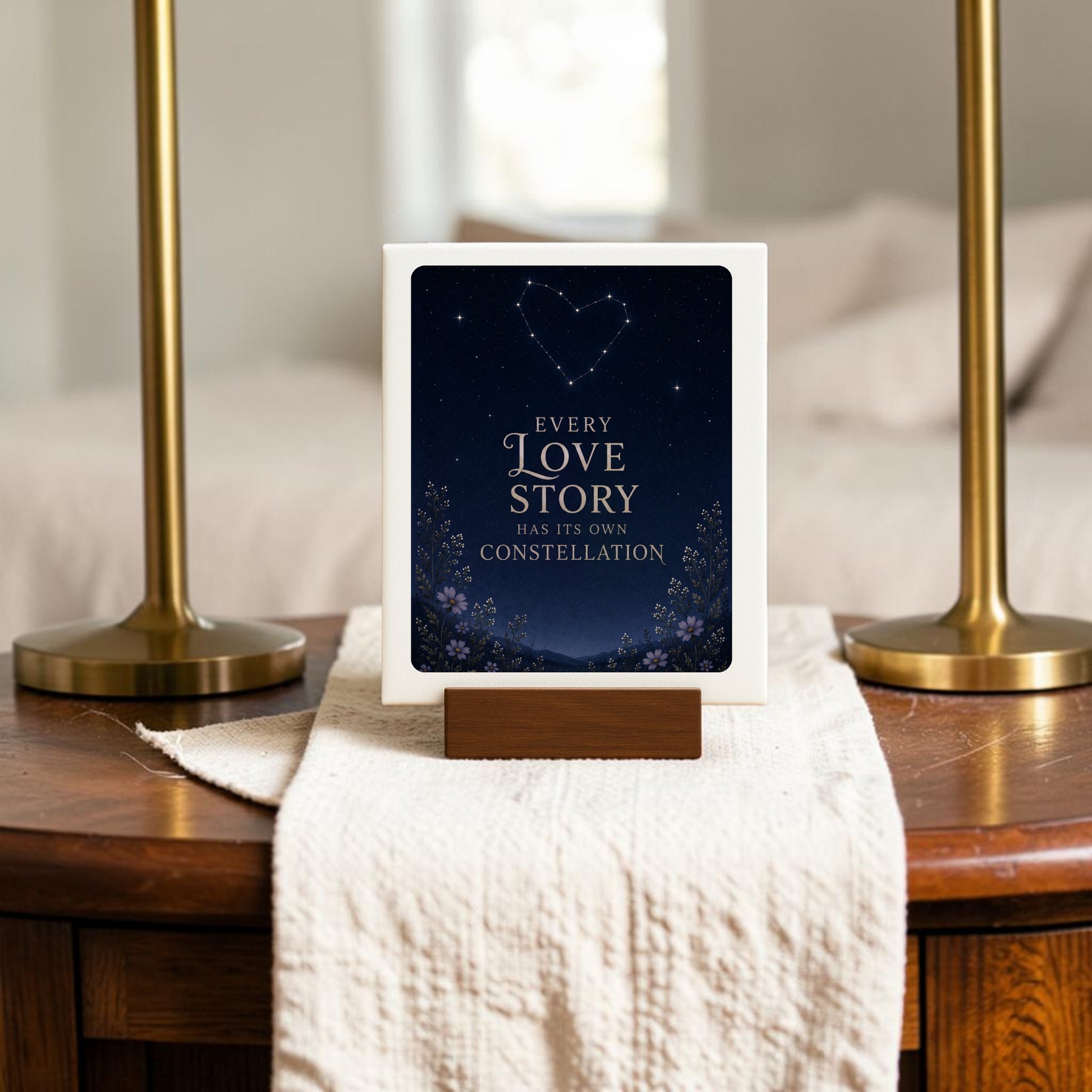 On a wooden table, the Every Love Story Constellation Mini Sign from Mirabilia Boutique features the words, “Every love story has its own constellation,” with a heart-shaped constellation above and delicate flowers below. Part of the enchanting Star & Solstice Collection, this celestial decor piece rests on a white cloth between two brass lamps.