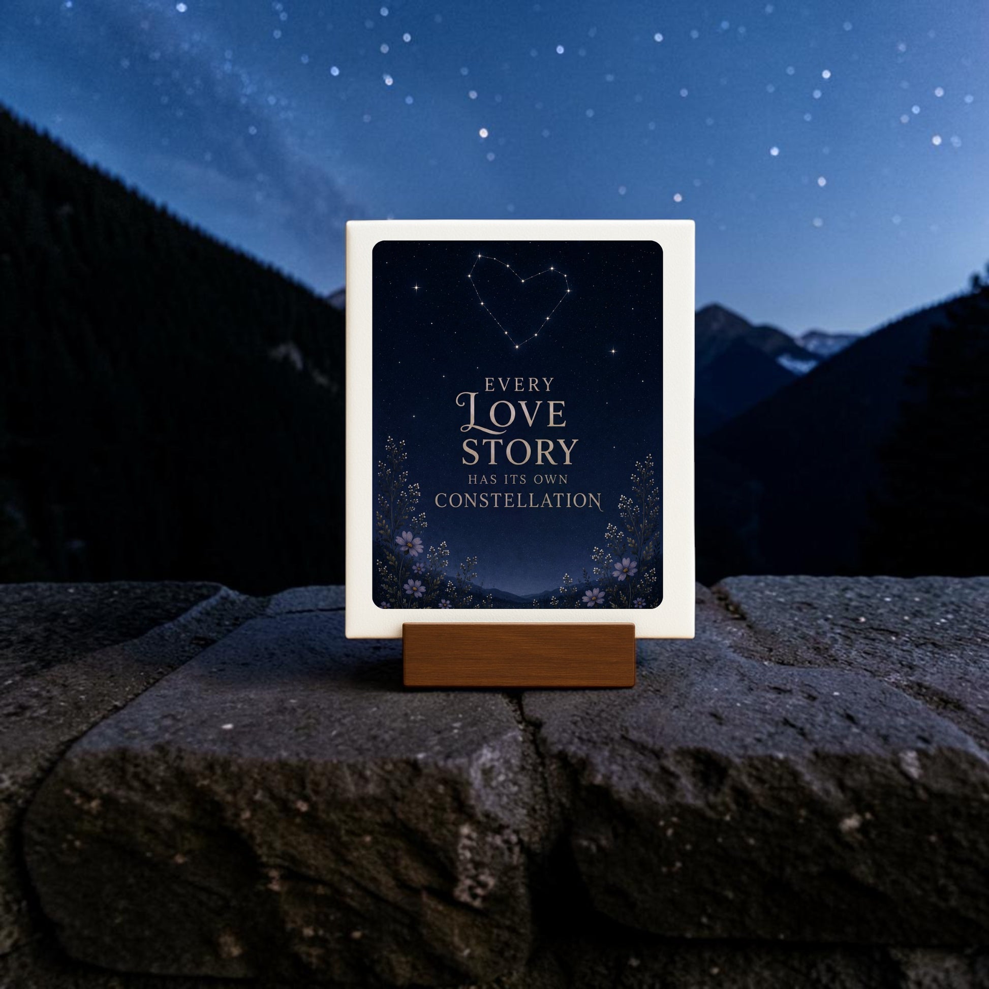 The Every Love Story Constellation Mini Sign by Mirabilia Boutique from the Star & Solstice Collection rests on a wooden stand, featuring the phrase “Every love story has its own constellation” alongside a heart-shaped constellation above mountains beneath a starry night sky—a charming piece of celestial decor.