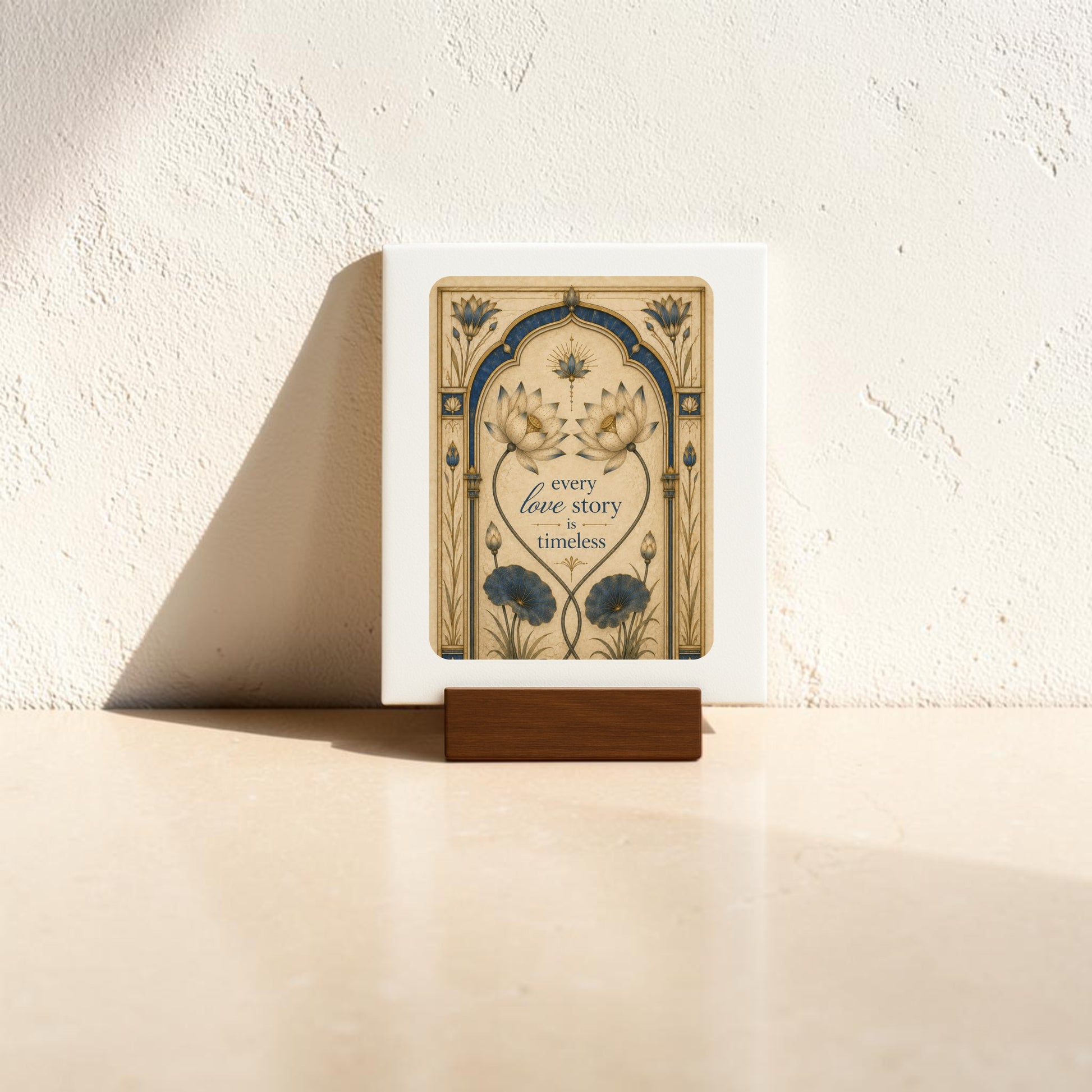 The Every Love Story Is Timeless | Lotus Mini Sign by Mirabilia Boutique features floral designs and displays the phrase “every love story is timeless.” This mini sign is showcased on a small wooden stand, set atop a light-colored surface against a textured wall, with sunlight casting gentle shadows.