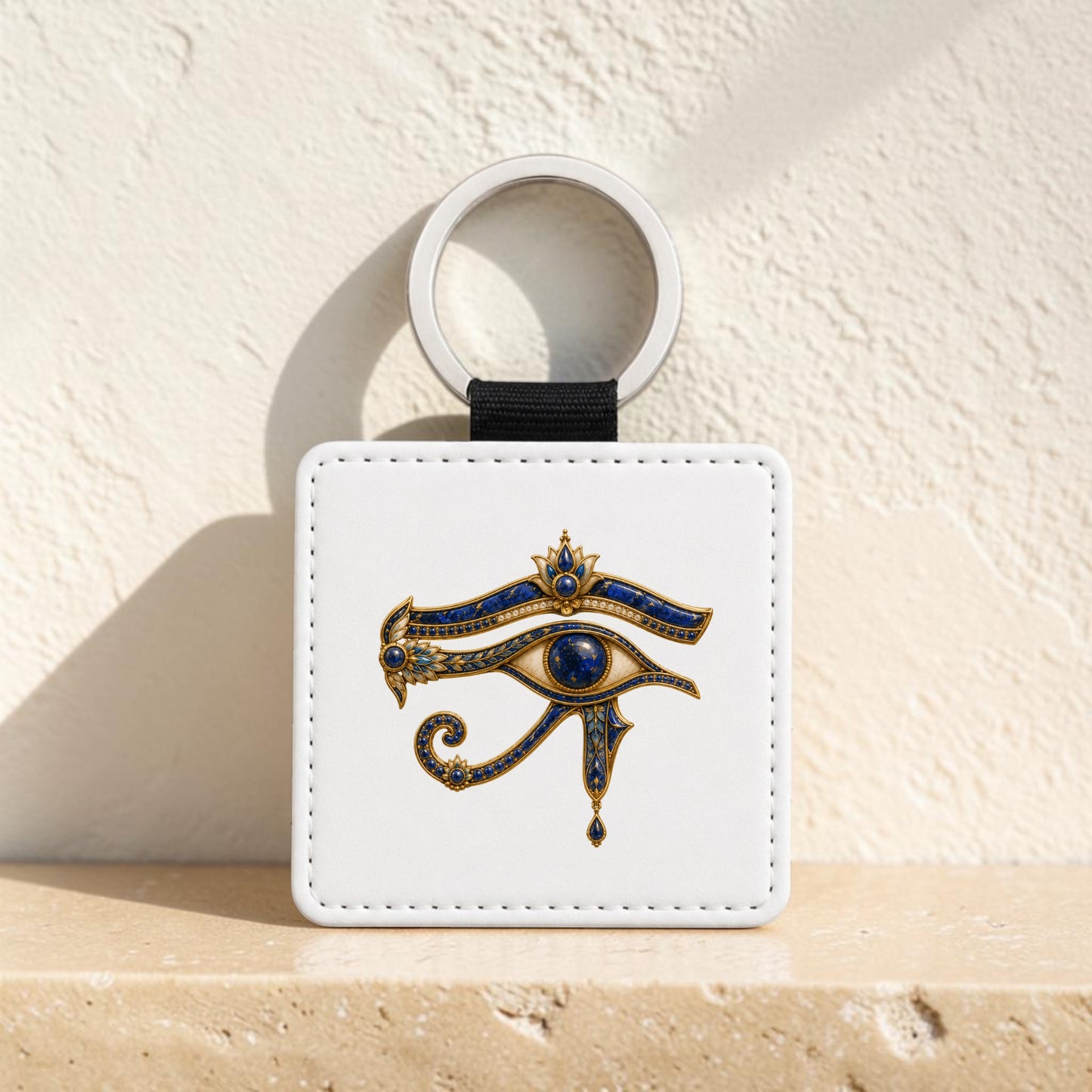 The Mirabilia Boutique Eye of Horus Keychain | Ancient Egyptian Keepsake, featuring a white square design adorned with a gold and lapis blue Eye of Horus symbol, stands upright on a beige stone surface against a textured light wall.