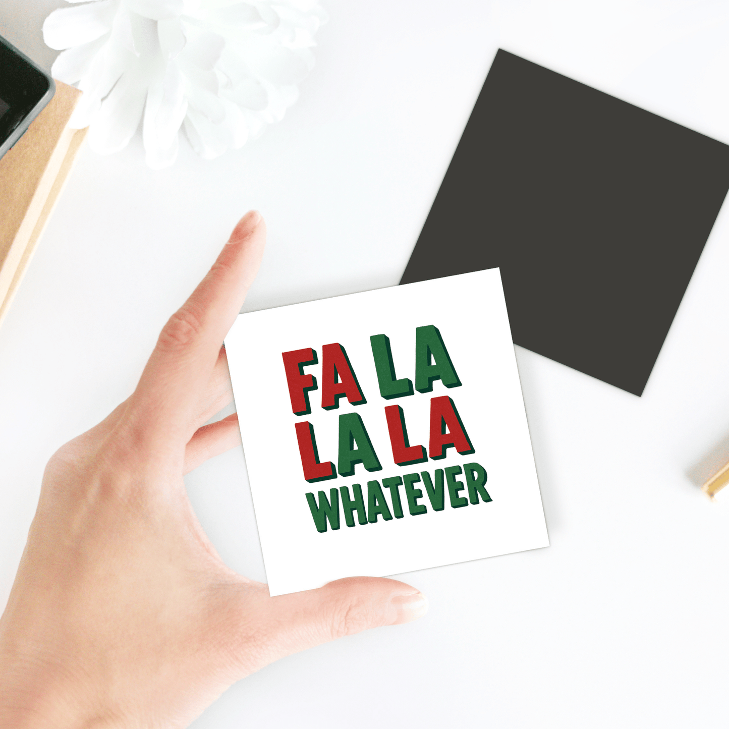 A hand holds the "Fa La La La Whatever Magnet" by Mirabilia Boutique, a funny Christmas gift idea. In the background, a black square and white flower rest on a white surface.