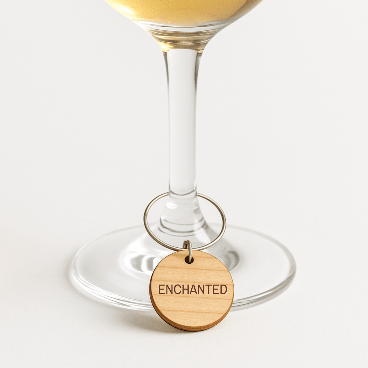A wine glass adorned with an "ENCHANTED" charm from the Mirabilia Boutique Enchanted Woodland Wine Charms Set hangs gracefully from its stem, resting on a white surface. This elegant wine charm set offers both style and practicality as delightful glass markers.