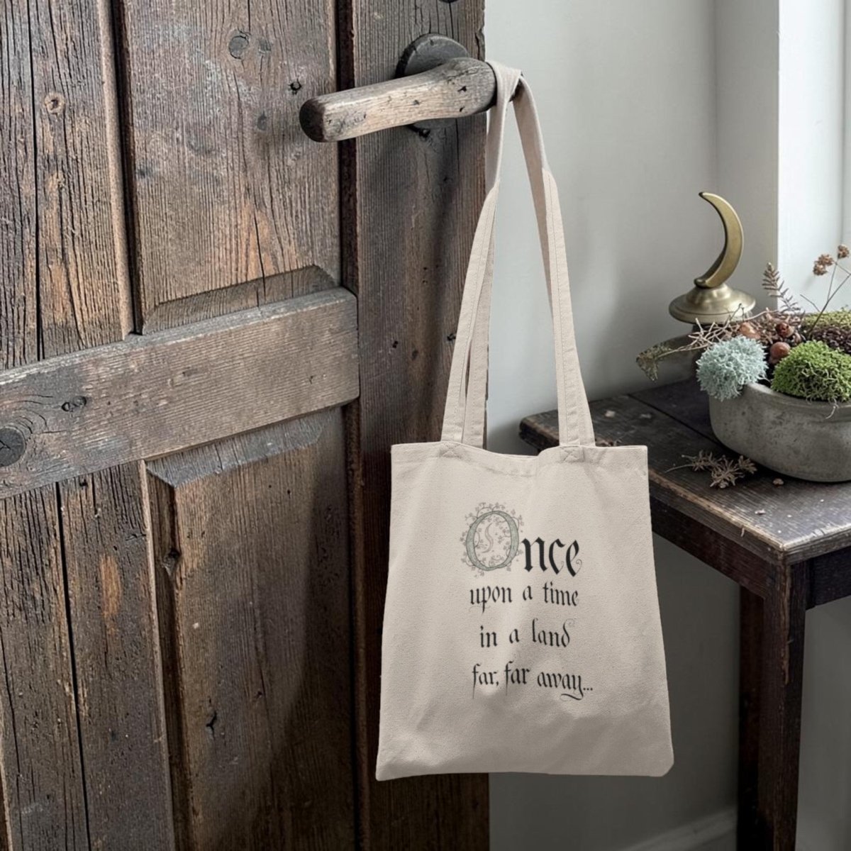 A beige Once Upon a Time Fairytale Tote Bag by Mirabilia Boutique, featuring the phrase "Once upon a time in a land far, far away...," hangs on a rustic wooden door. Nearby, a small table displays a crescent moon sculpture and a bowl filled with greenery and dried flowers.