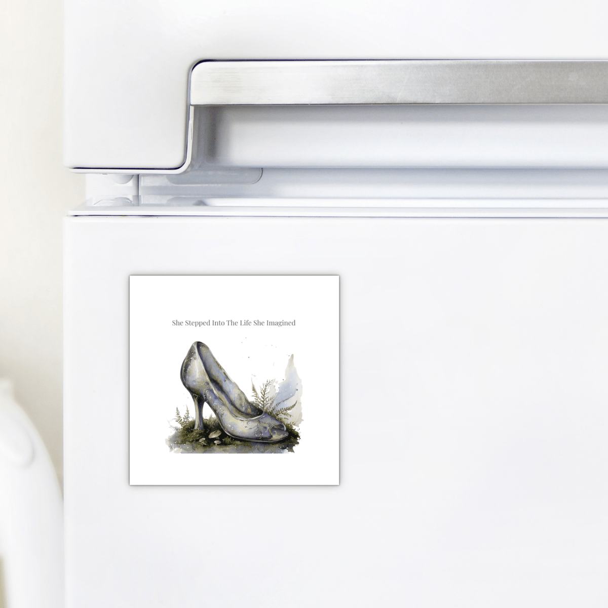 The She Stepped Into the Life She Imagined Magnet by Mirabilia Boutique is a square inspirational refrigerator magnet, shown on a white fridge, featuring a watercolor illustration of a high heel shoe embellished with a butterfly and greenery—ideal for fairytale decor. The text above reads, “She Stepped Into The Life She Imagined.”.