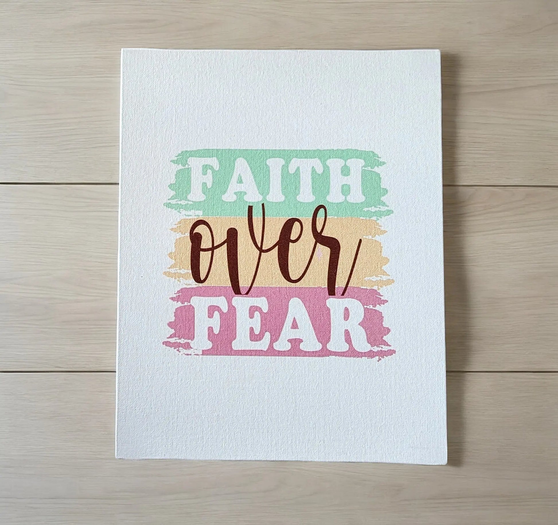 The Faith Over Fear Canvas Art by Mirabilia Boutique features a motivational phrase in vibrant pastel hues on a light wooden background. "Faith" and "Fear" are presented in bold uppercase letters, with "over" elegantly written in cursive, making it an ideal inspirational gift.