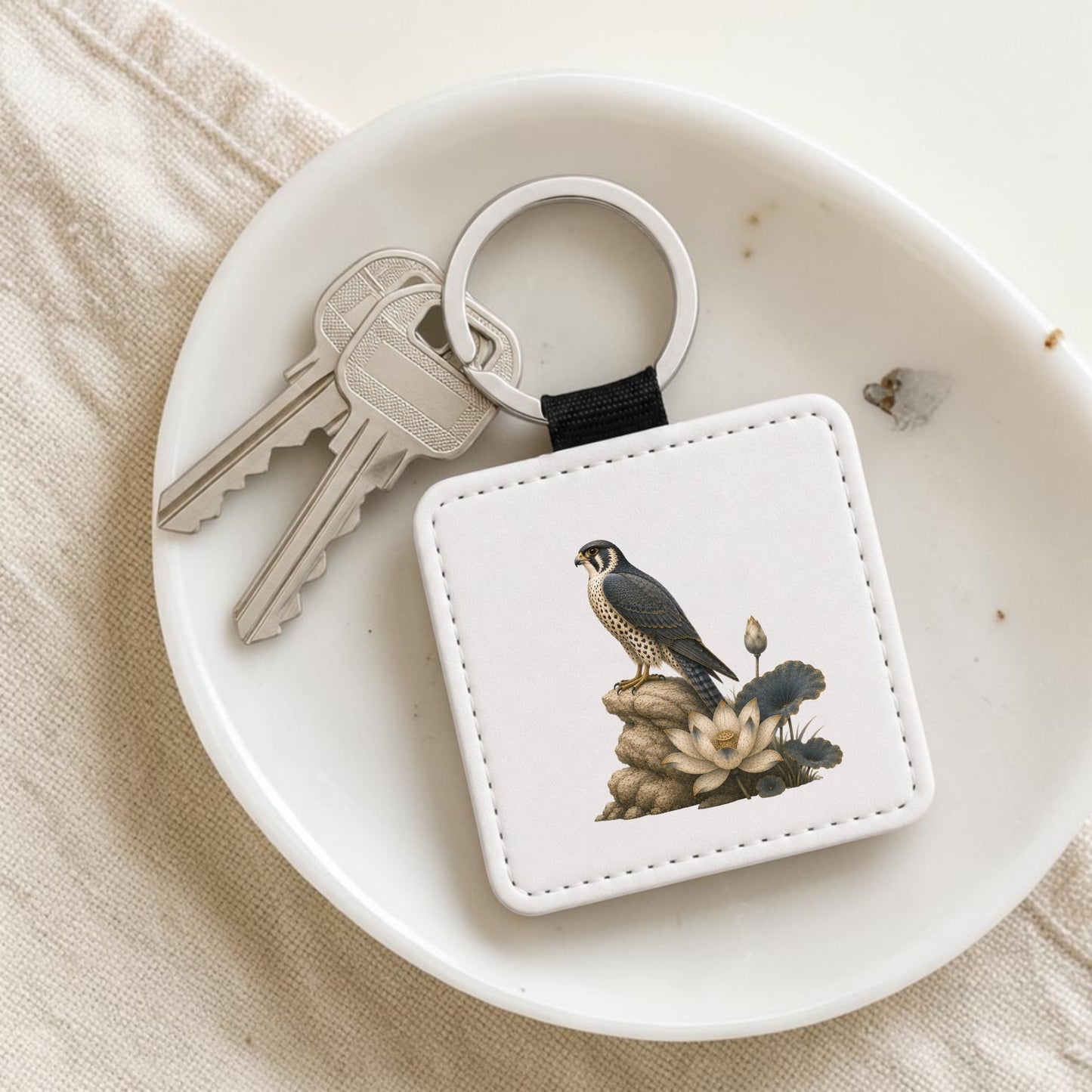 The Falcon & Lotus Keychain | Ancient Egyptian Art by Mirabilia Boutique—a white keychain featuring a falcon and lotus inspired by ancient Egyptian art—rests on a dish beside two silver keys.
