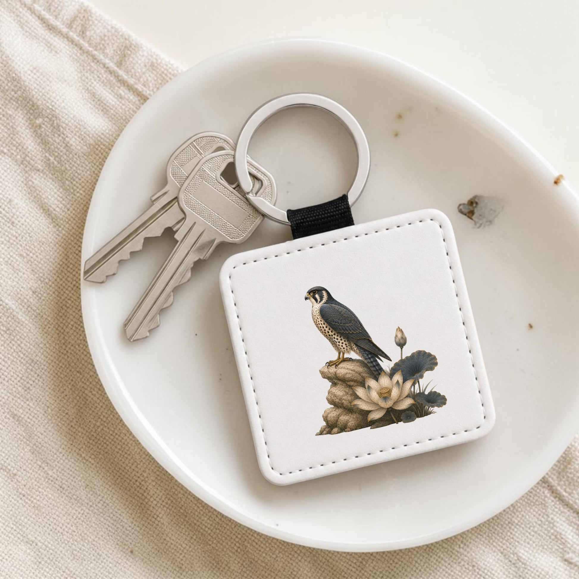 The Falcon & Lotus Keychain | Ancient Egyptian Art by Mirabilia Boutique—a white keychain featuring a falcon and lotus inspired by ancient Egyptian art—rests on a dish beside two silver keys.