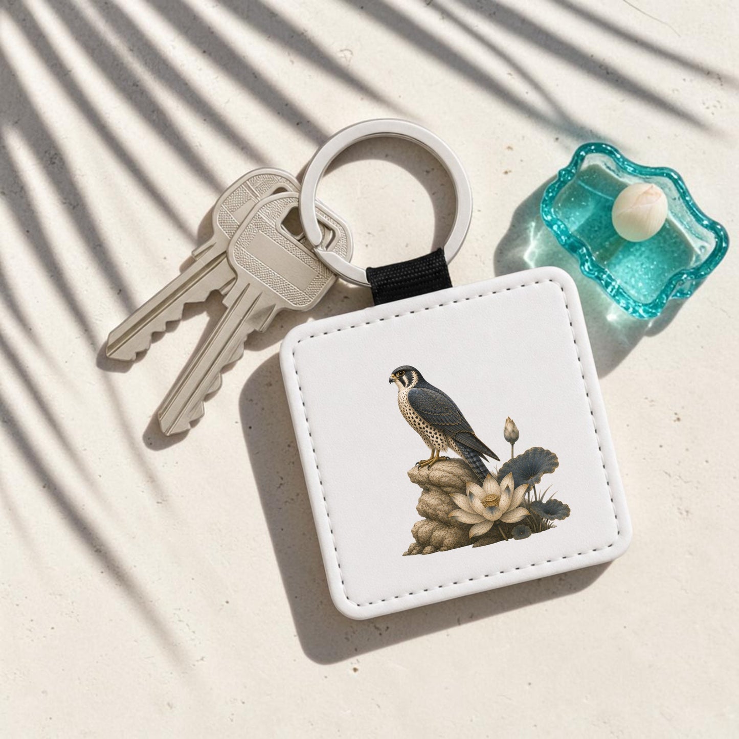 The Falcon & Lotus Keychain | Ancient Egyptian Art by Mirabilia Boutique—a square keychain adorned with a bird motif inspired by ancient Egyptian art—rests on a white surface next to two keys and a small aqua dish holding a marble, while leaf shadows fall across the scene.