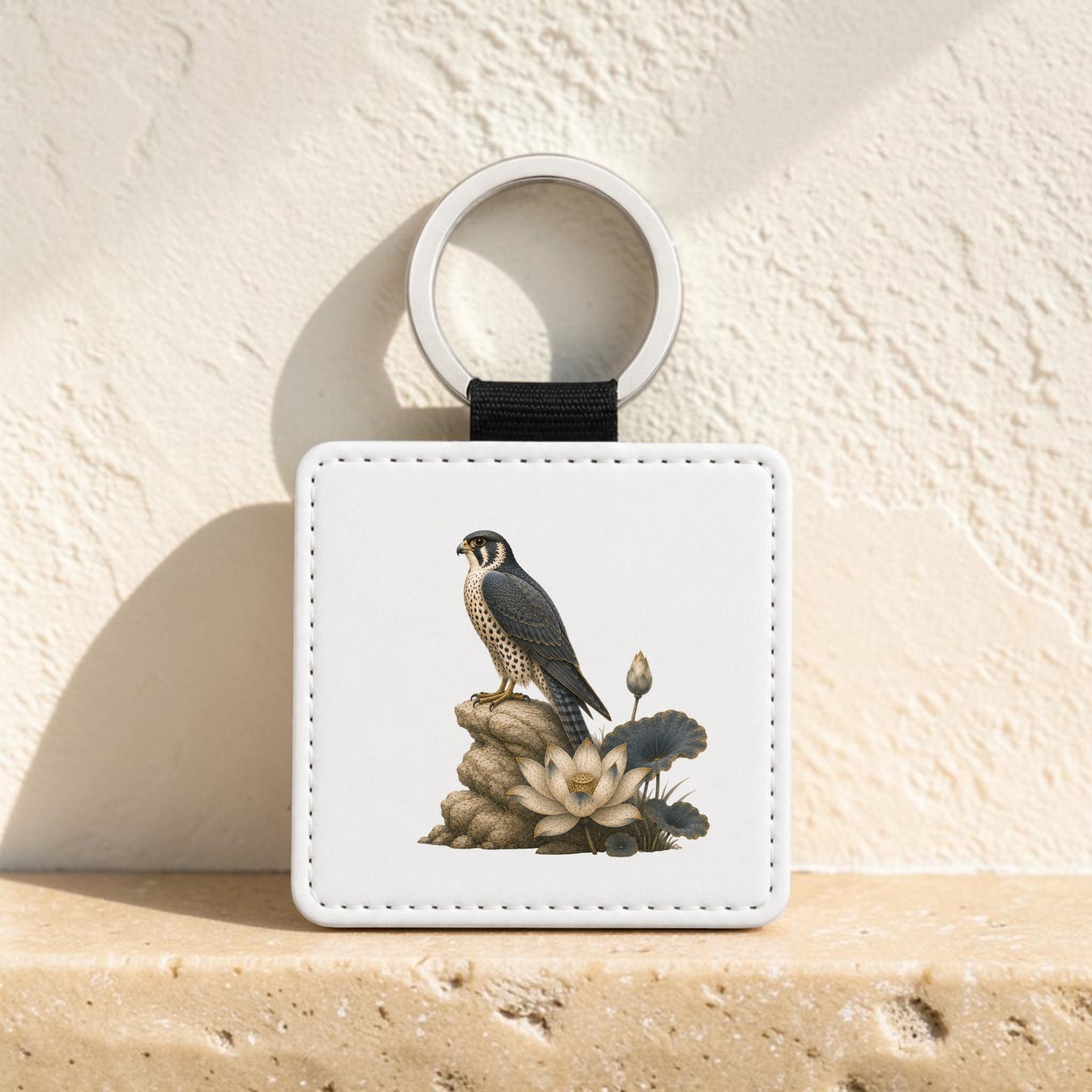 The Falcon & Lotus Keychain | Ancient Egyptian Art by Mirabilia Boutique is a square white keychain with a metal ring, featuring an illustration of a falcon and lotus perched on rocks and white flowers, displayed against a textured beige wall.