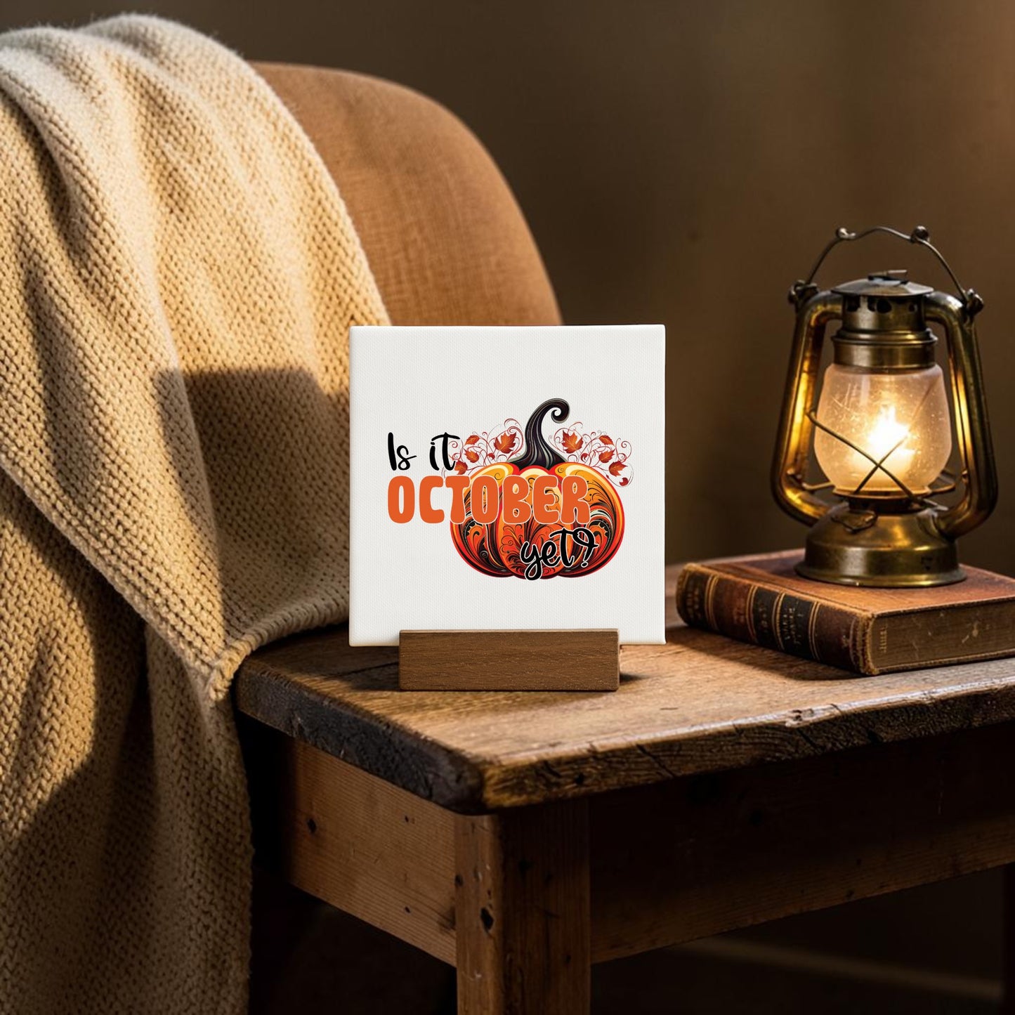 The Is It October Yet? Mini Sign by Mirabilia Boutique, featuring a pumpkin design, rests on a wooden easel on a rustic table beside a lantern, books, and a cream knit blanket draped over a chair.