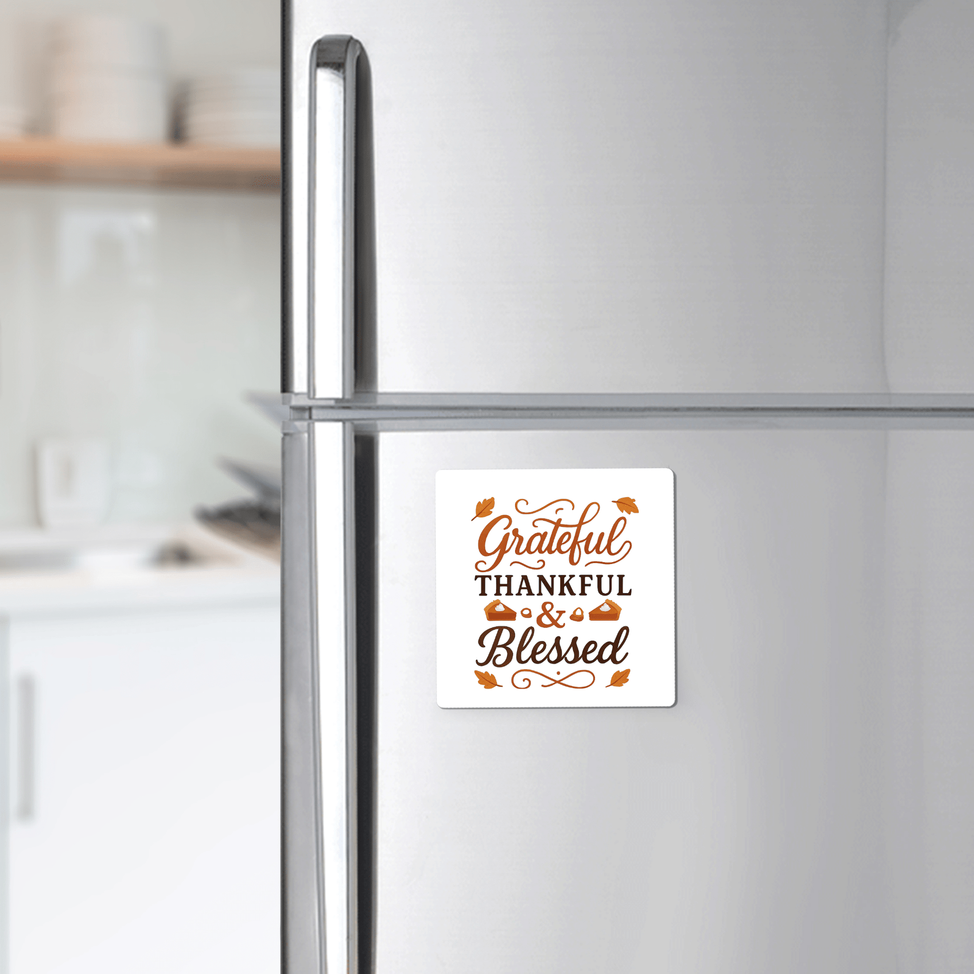 A silver fridge displays the Mirabilia Boutique "Grateful Thankful Blessed Magnet" in brown and orange with leafy accents—an ideal fall or Thanksgiving decor piece. A blurred kitchen is visible in the background.