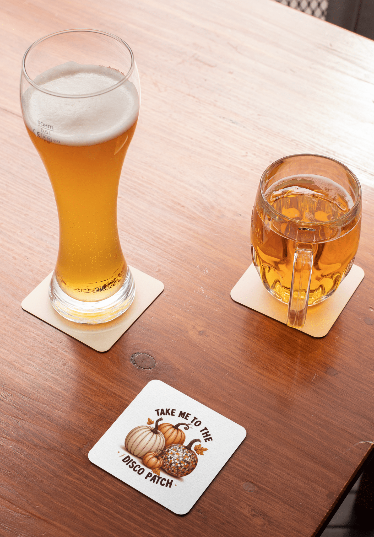 A tall glass of beer and a mug of cider sit on coasters atop a wooden table. A third coaster from Mirabilia Boutique’s Disco Patch Pumpkin Coaster Set, featuring retro pumpkin illustrations, completes the scene.