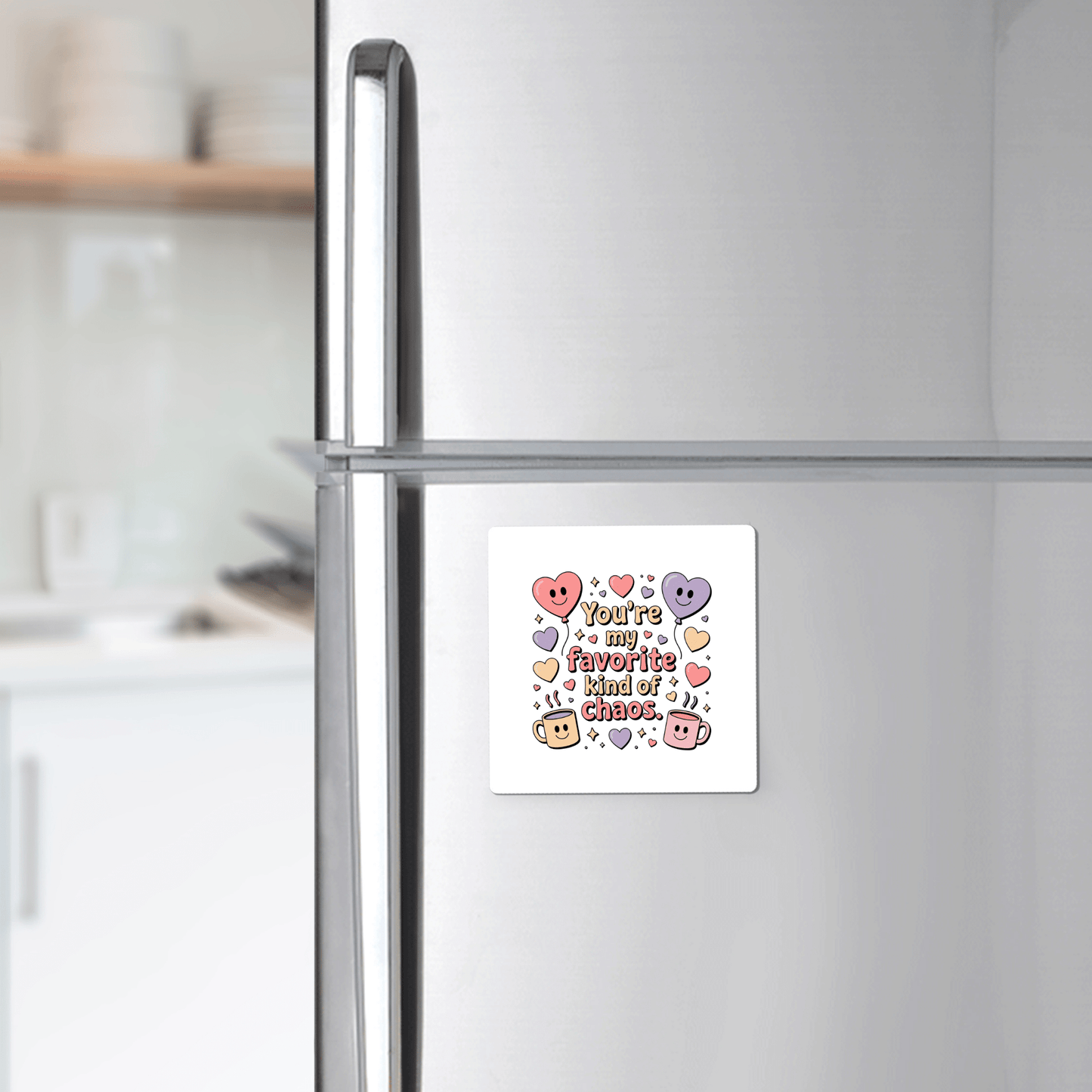 A Mirabilia Boutique “Favorite Kind of Chaos Gift for Friends” fridge magnet with colorful lettering and cute cartoon hearts and stars decorates a silver refrigerator in a softly blurred kitchen. Perfect for adding charm and humor to any space.