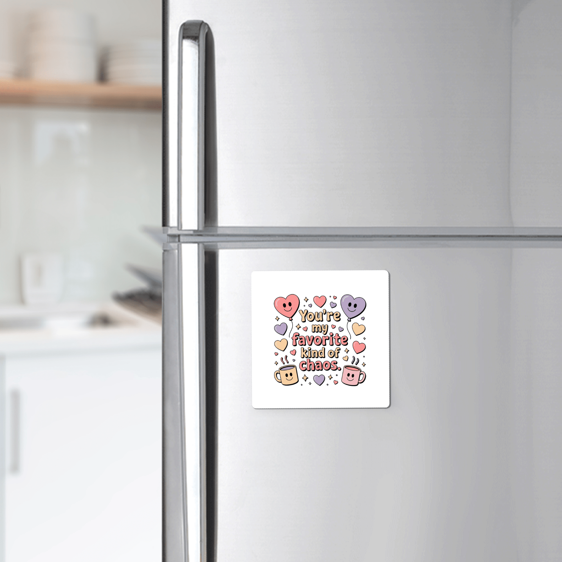 A Mirabilia Boutique “Favorite Kind of Chaos Gift for Friends” fridge magnet with colorful lettering and cute cartoon hearts and stars decorates a silver refrigerator in a softly blurred kitchen. Perfect for adding charm and humor to any space.