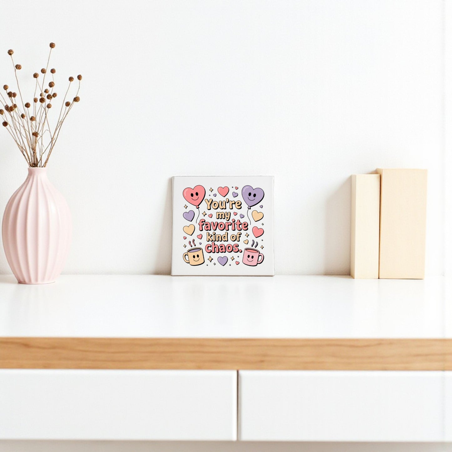 A minimal white shelf shows Mirabilia Boutique's "Favorite Kind of Chaos" canvas—a funny Valentine’s or Galentine’s gift—featuring colorful hearts, a pink vase with dried branches, and two beige books for charming decor.