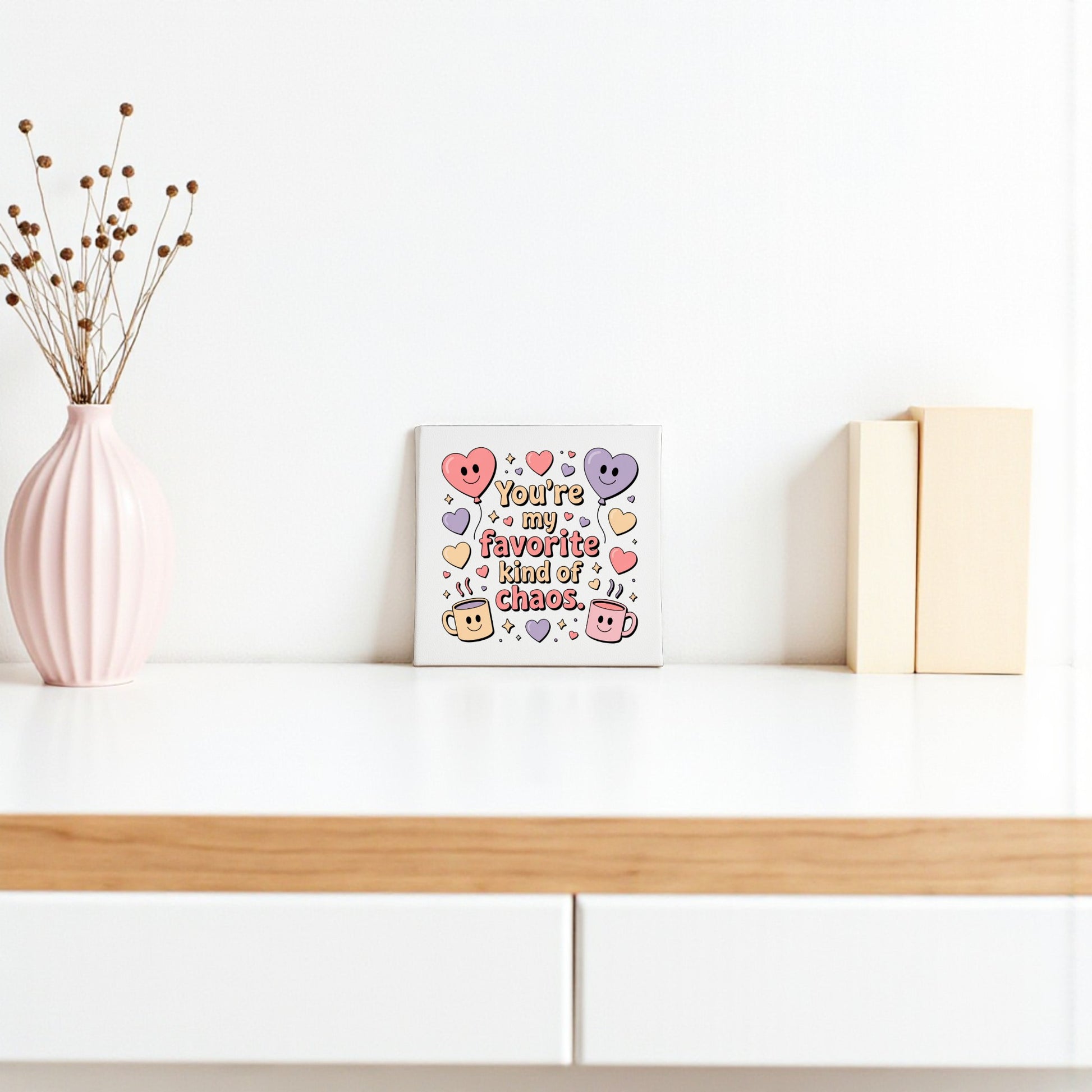 A minimal white shelf shows Mirabilia Boutique's "Favorite Kind of Chaos" canvas—a funny Valentine’s or Galentine’s gift—featuring colorful hearts, a pink vase with dried branches, and two beige books for charming decor.
