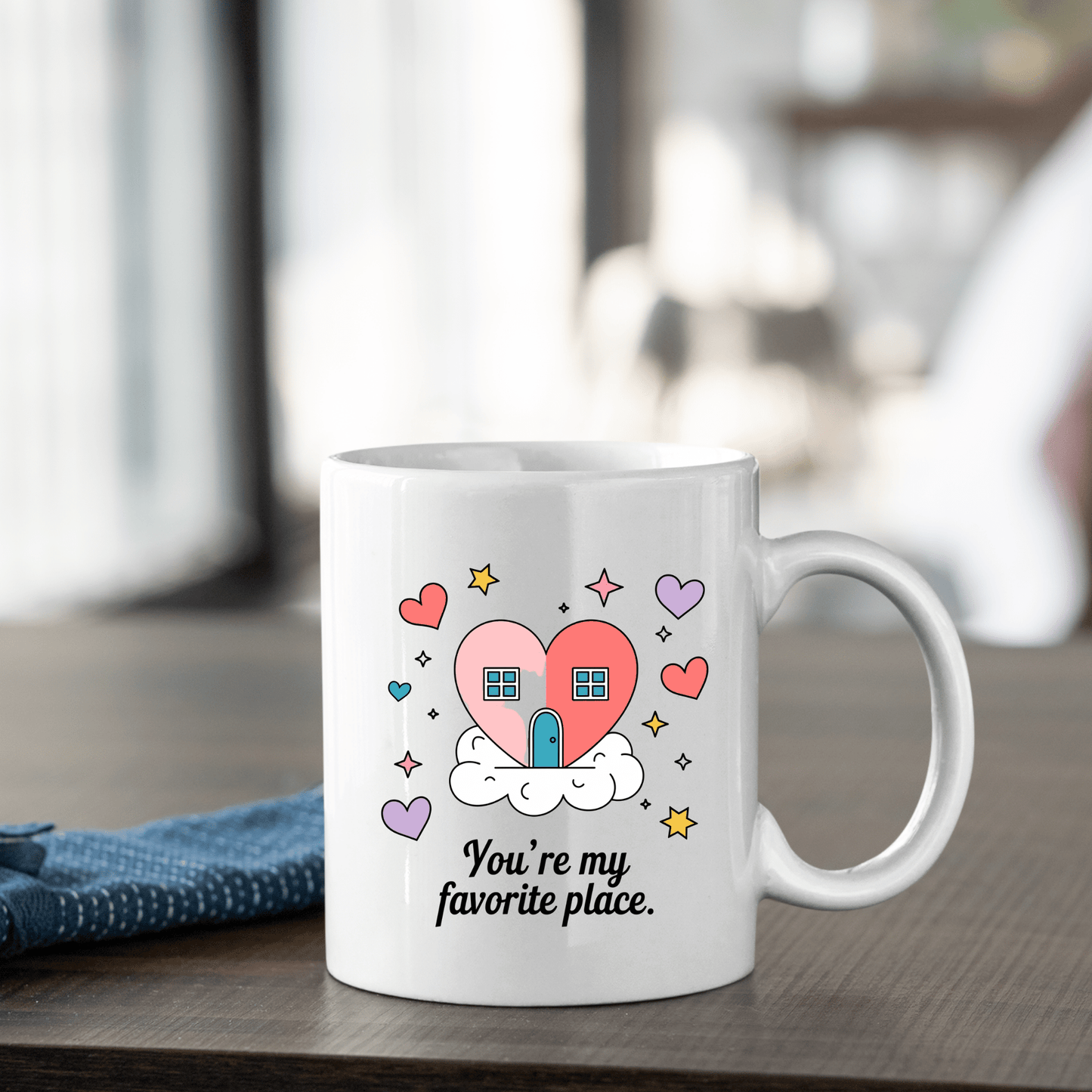 The Heart House Mug by Mirabilia Boutique features a pink heart-shaped house on a cloud with colorful hearts and stars, plus the phrase "You're my favorite place." A blue cloth rests beside this cozy gift mug.