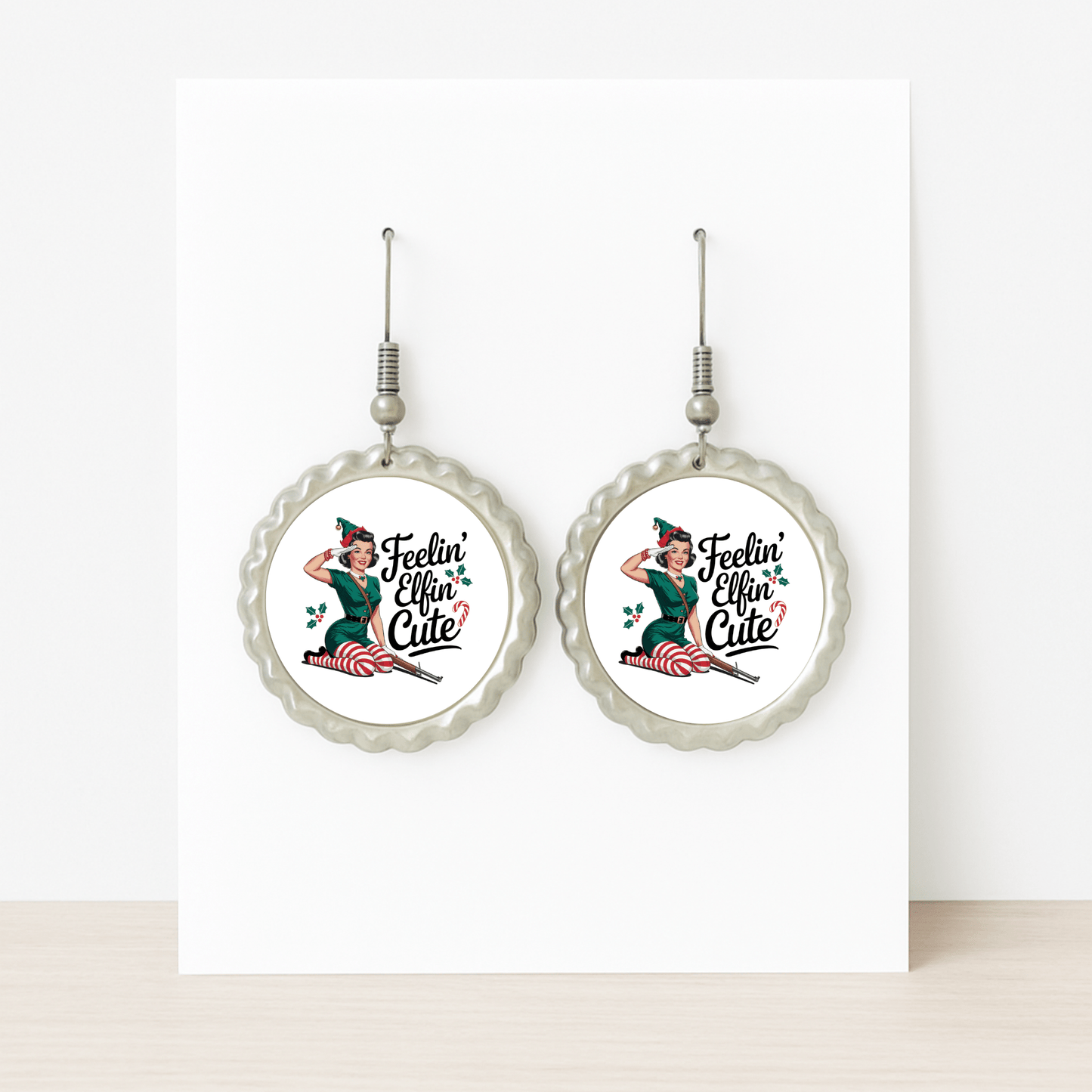 The Feelin’ Elfin’ Cute Earrings by Mirabilia Boutique are round, silver retro Christmas earrings with a pin-up girl and holly leaves—funny holiday jewelry for women, displayed on a white card and perfect for festive occasions.