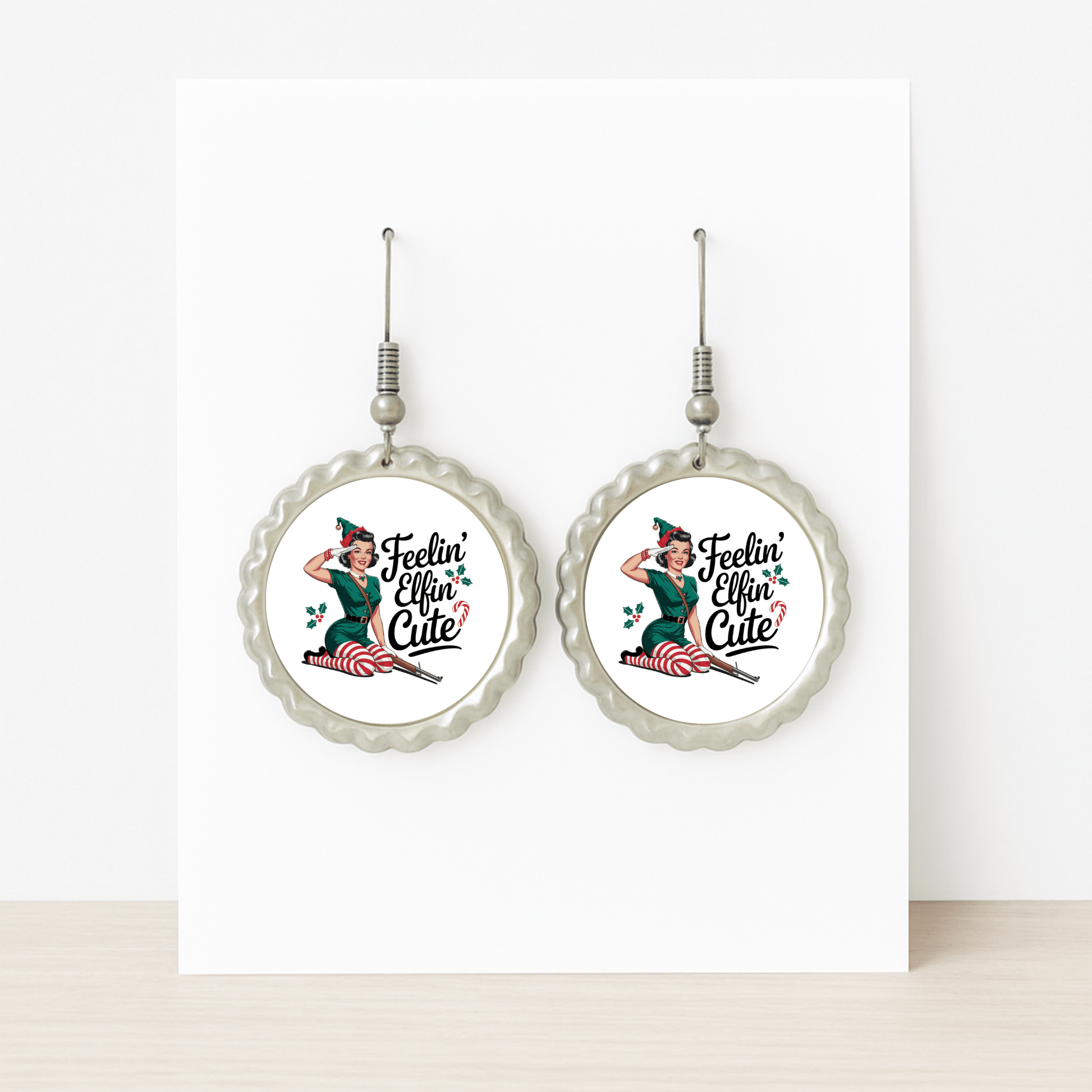 The Feelin’ Elfin’ Cute Earrings by Mirabilia Boutique are round, silver retro Christmas earrings with a pin-up girl and holly leaves—funny holiday jewelry for women, displayed on a white card and perfect for festive occasions.