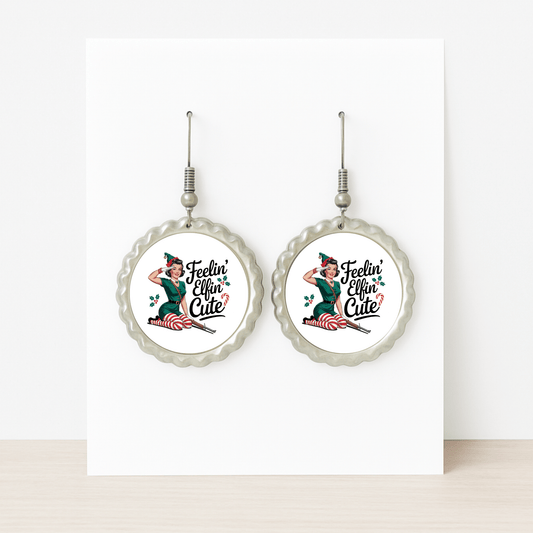 The Feelin’ Elfin’ Cute Earrings by Mirabilia Boutique are round, silver retro Christmas earrings with a pin-up girl and holly leaves—funny holiday jewelry for women, displayed on a white card and perfect for festive occasions.