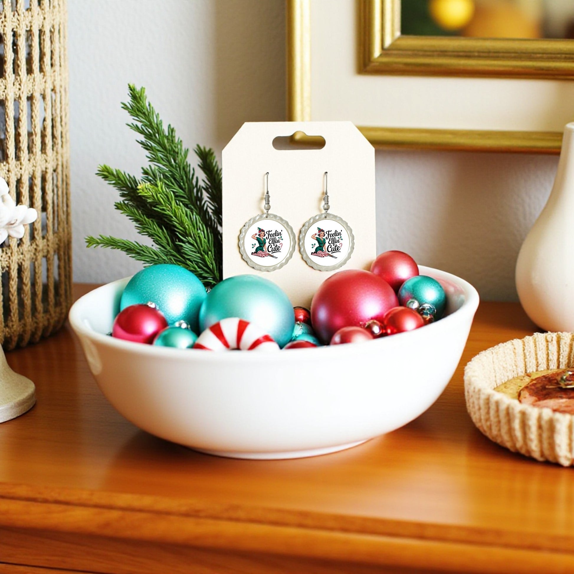 A white bowl of colorful Christmas ornaments and greenery sits on a wooden table. Featured among the decor are Mirabilia Boutique’s Feelin’ Elfin’ Cute Earrings—a playful, funny holiday jewelry gift for women, displayed on their branded card.
