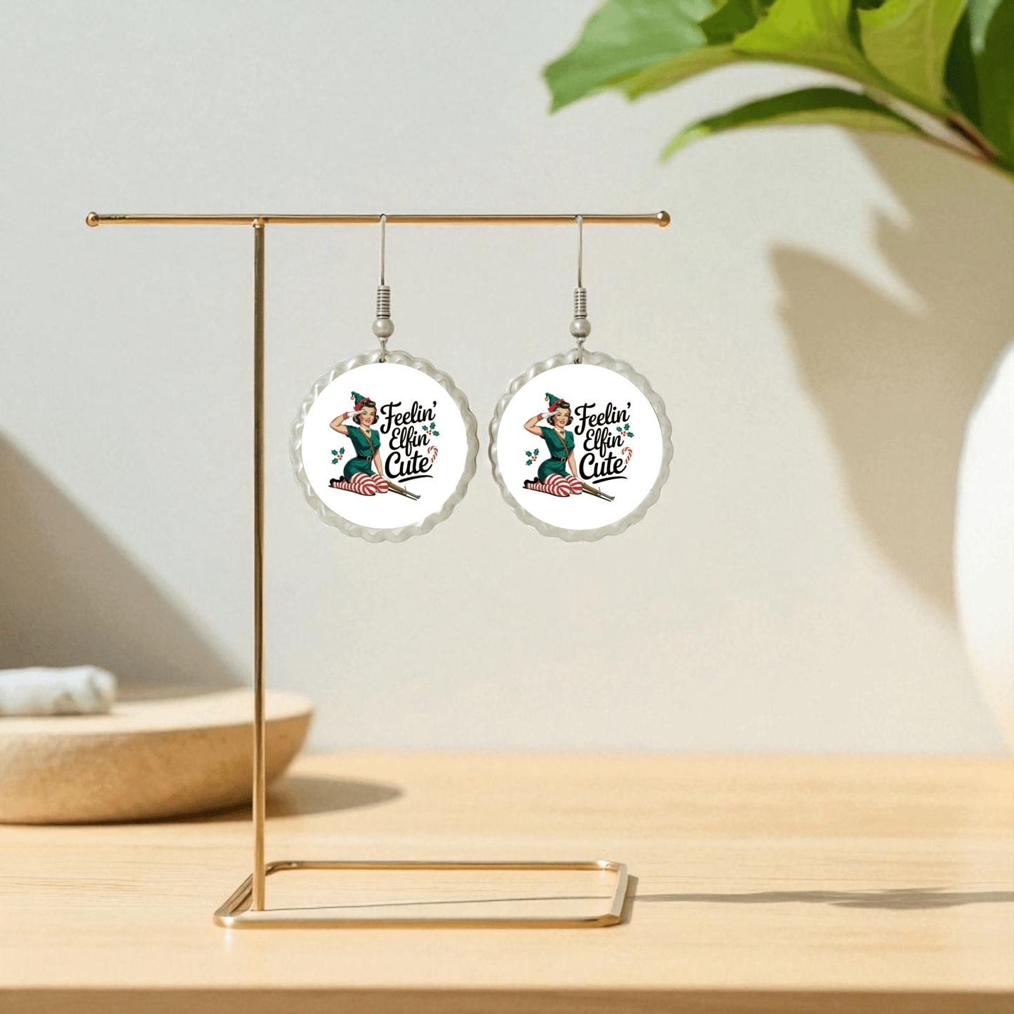 Feelin’ Elfin’ Cute Earrings by Mirabilia Boutique, displayed on a gold stand, feature a festive retro pin-up mermaid and playful holiday text. Background shows a blurred plant and neutral decor on light wood. Funny holiday jewelry for women.