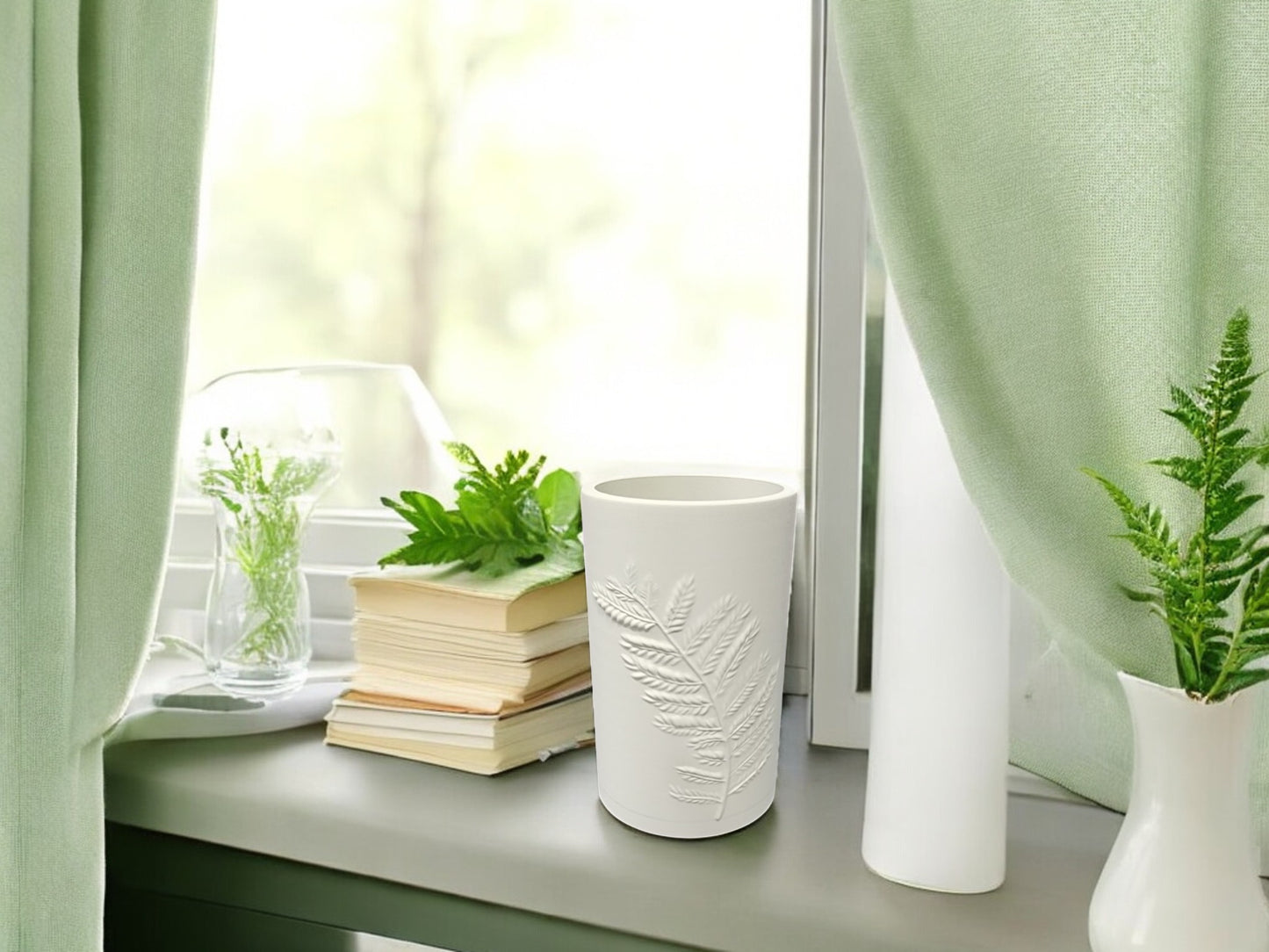 A Mirabilia Boutique Fern Emboss Vase in Soft Green or Matte Beige sits on a windowsill beside books, a glass with green plants, and light green curtains—an elegant touch for minimalist decor.