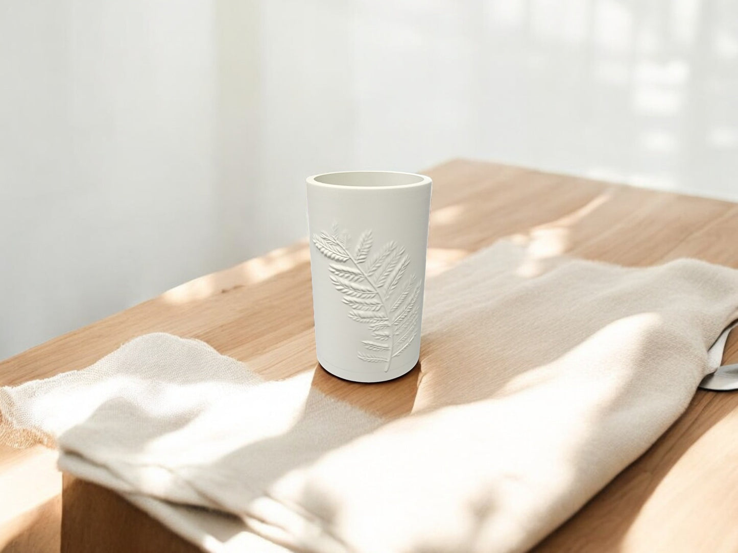 The Fern Emboss Vase by Mirabilia Boutique, available in soft green or matte beige, rests on a light wooden table beside a folded beige cloth in gentle sunlight—a perfect accent for minimalist decor.