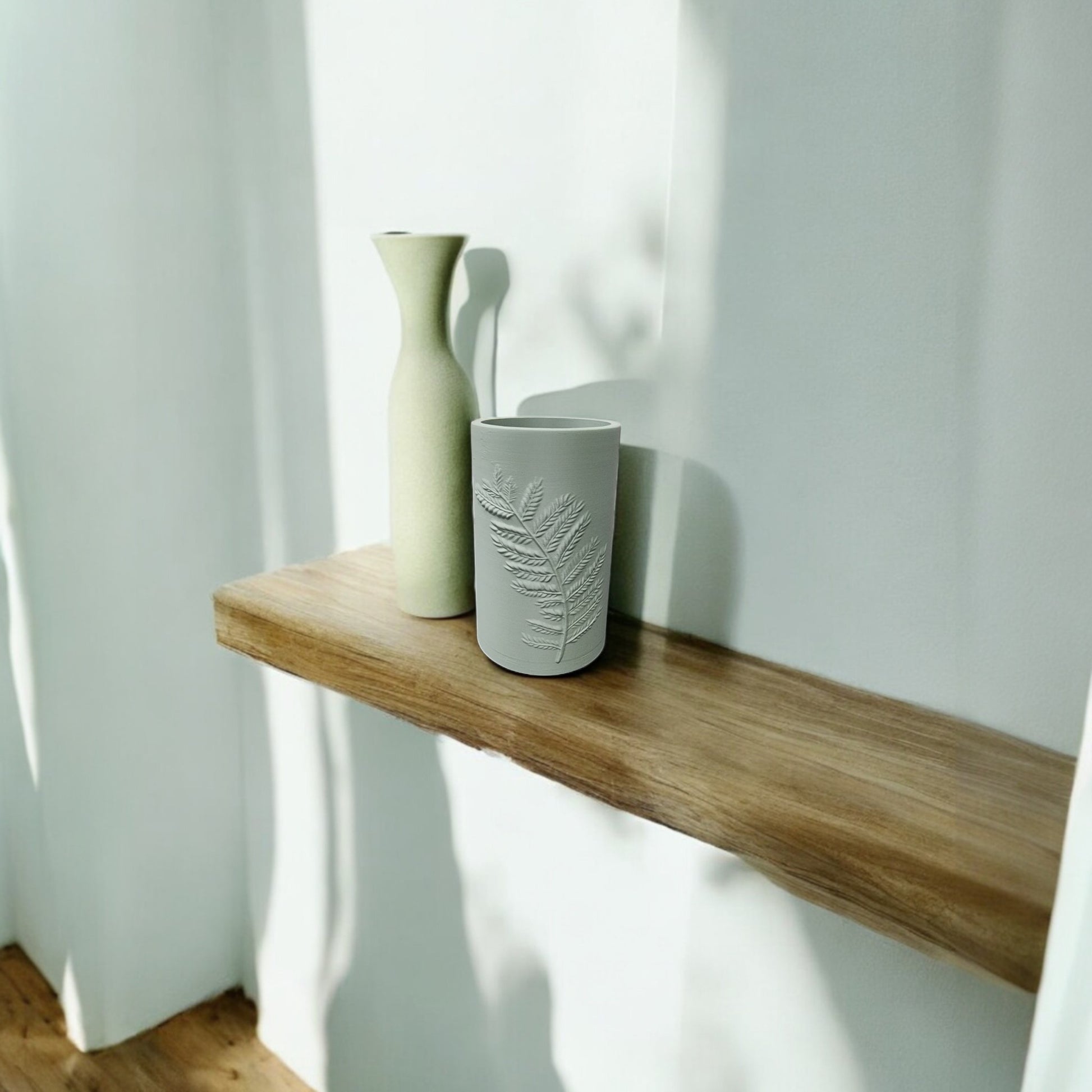 A minimalist wooden shelf holds a tall, narrow beige vase and Mirabilia Boutique’s Fern Emboss Vase in matte beige, both ideal for elevating minimalist decor, set against a white wall in natural sunlight.