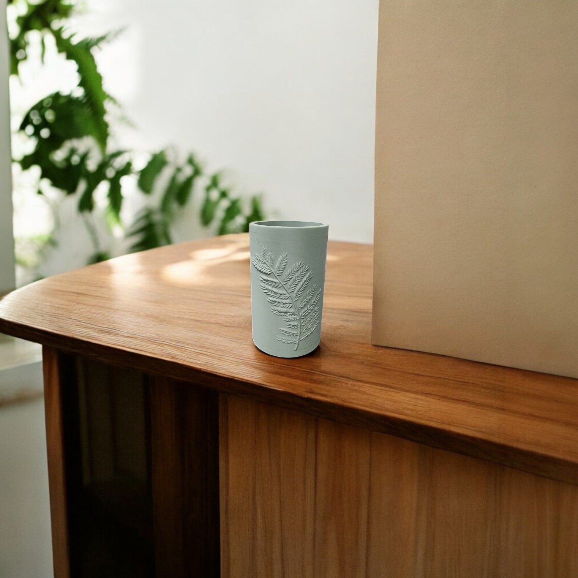 A Fern Emboss Vase in matte beige by Mirabilia Boutique sits on a wooden table with a light blue cup, surrounded by green plants and sunlight, creating a serene minimalist decor scene.
