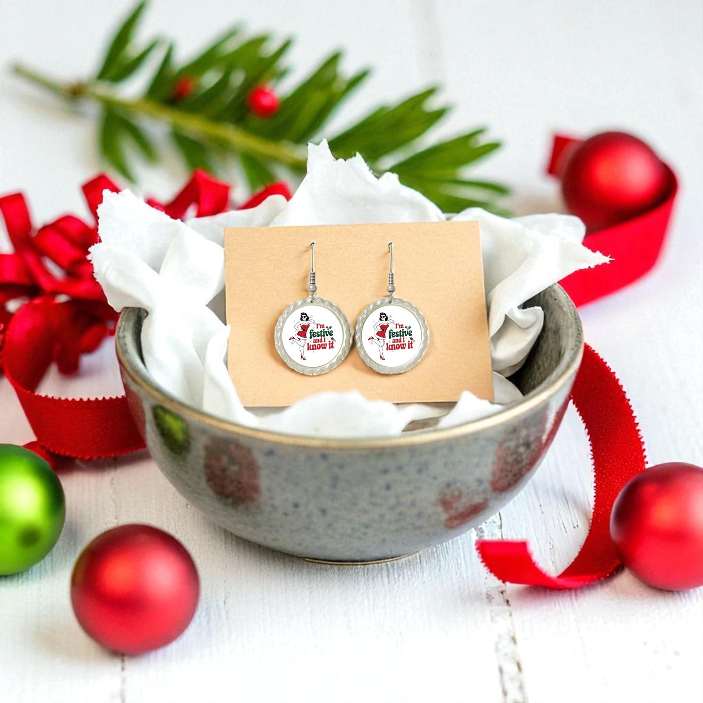 The Festive and I Know It Earrings by Mirabilia Boutique are displayed in a ceramic bowl with tissue paper, surrounded by Christmas ornaments, red ribbon, and pine sprigs on a white wooden surface. Retro holiday style for the season!.