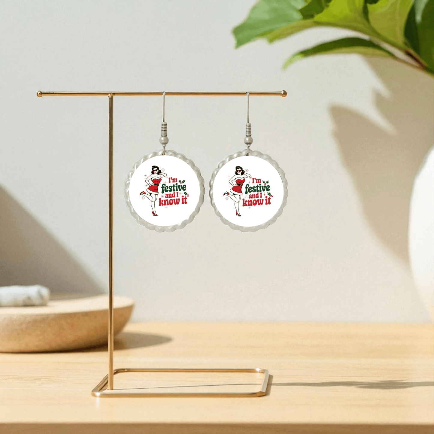 The Festive and I Know It Earrings by Mirabilia Boutique feature retro pin-up women in Santa hats with the phrase "I'm festive and I know it," shown on a gold stand with a plant in the background.