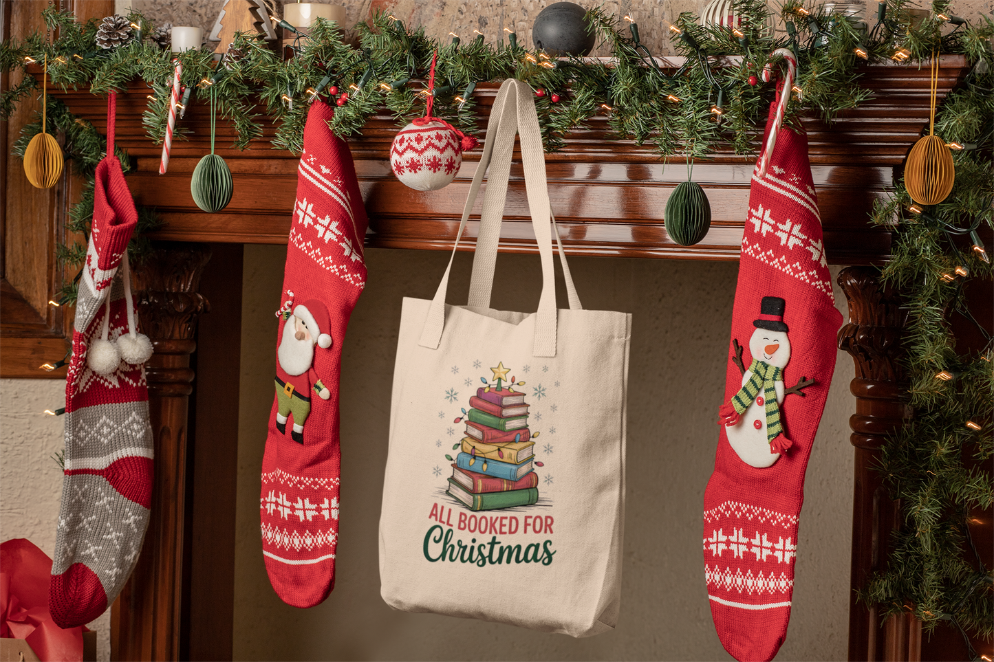 A festive mantel with garland, ornaments, three red stockings, and the Mirabilia Boutique "All Booked for Christmas Book Stack Tote Bag"—a perfect gift for book lovers.