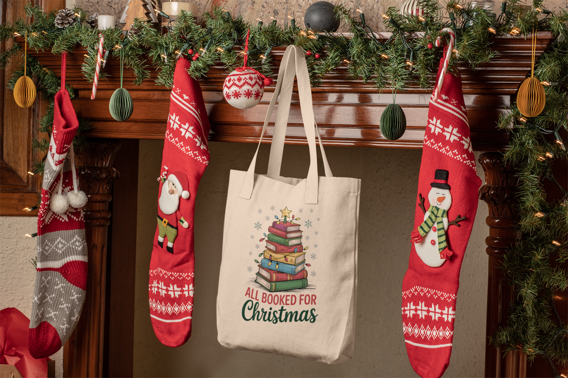 A festive mantel with garland, ornaments, three red stockings, and the Mirabilia Boutique "All Booked for Christmas Book Stack Tote Bag"—a perfect gift for book lovers.
