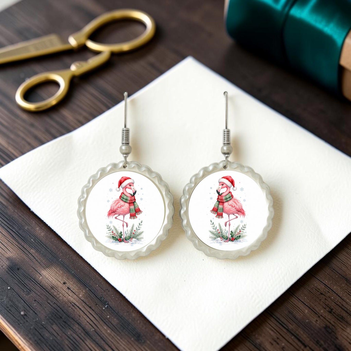 Add a festive touch to your holiday style with the Festive Flamingo Earrings | Winter Bird Jewelry by Mirabilia Boutique—handcrafted earrings depicting flamingos in Santa hats and scarves, displayed on a white napkin alongside gold scissors and a green-wrapped gift box atop a wooden surface. Celebrate the season in style with this unique holiday jewelry!.