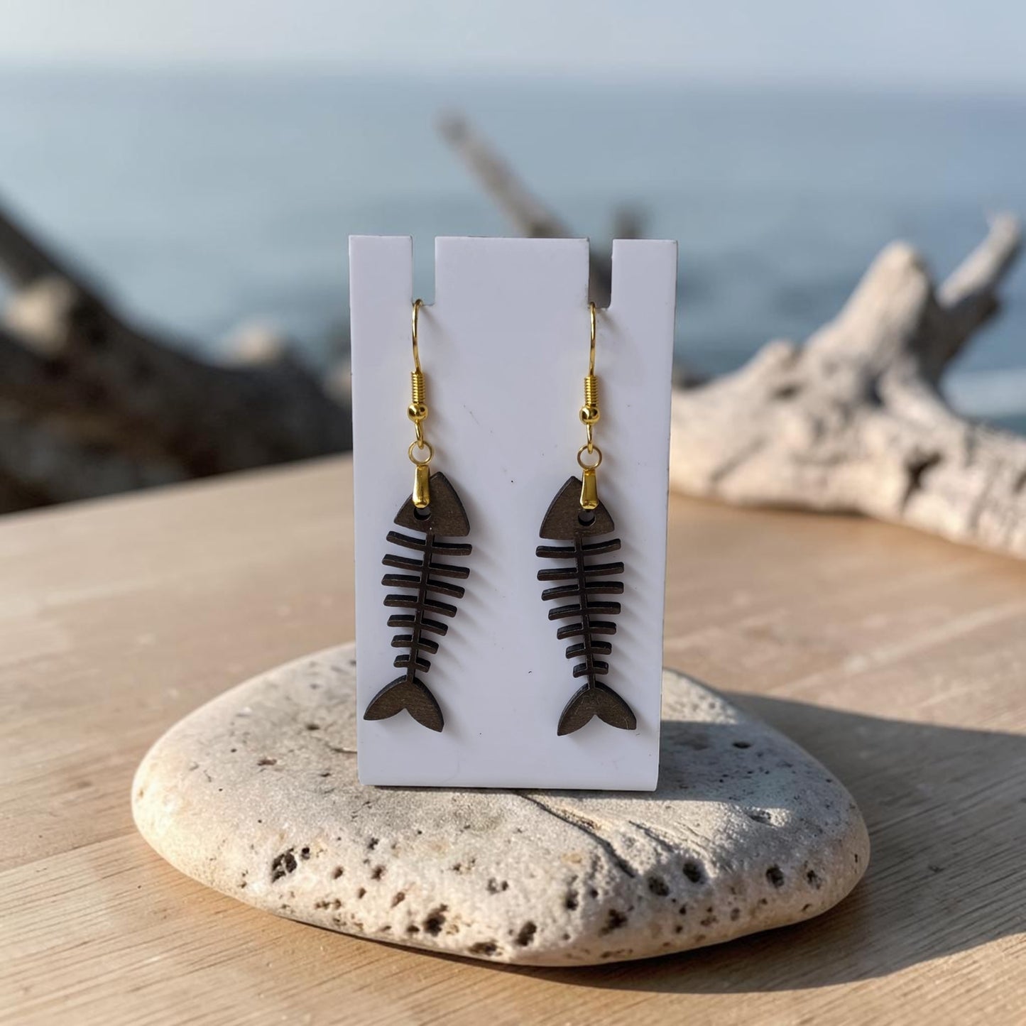 Fishbone Earrings | Lasercut Walnut Wood by Mirabilia Boutique, featuring gold hooks, are shown on a white card atop a stone and wood surface with a blurred seaside backdrop—ideal for fans of unique coastal jewelry.
