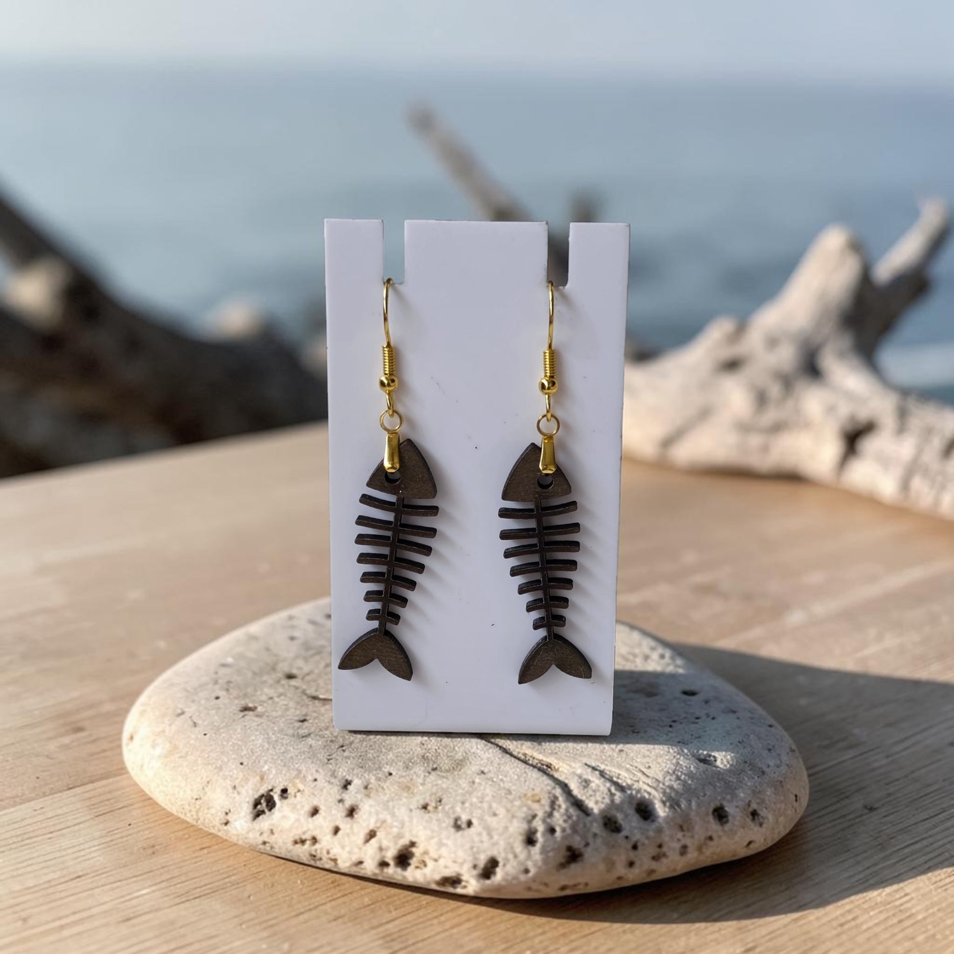 Fishbone Earrings | Lasercut Walnut Wood by Mirabilia Boutique, featuring gold hooks, are shown on a white card atop a stone and wood surface with a blurred seaside backdrop—ideal for fans of unique coastal jewelry.