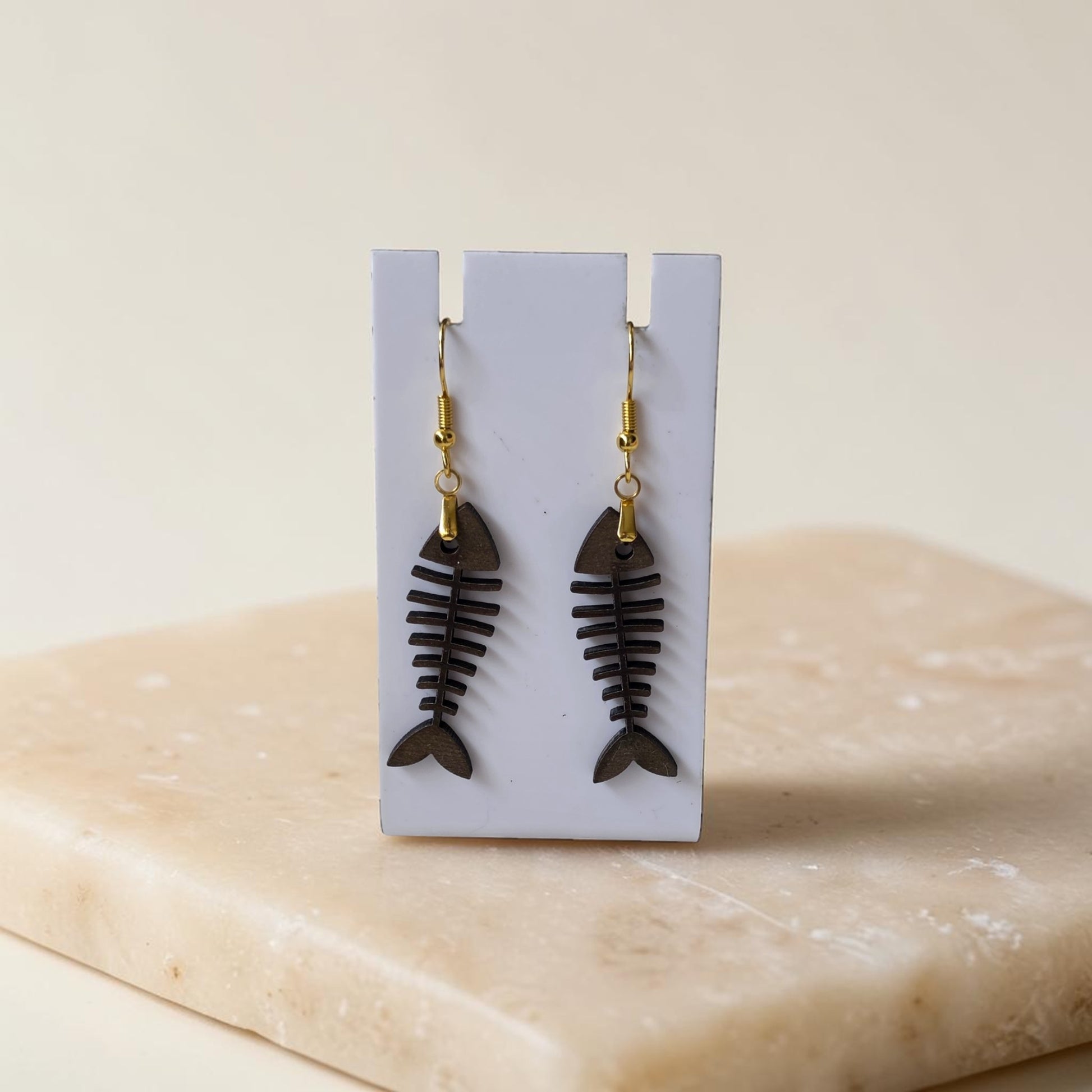 Fishbone Earrings by Mirabilia Boutique, crafted from lasercut walnut wood with gold hooks, are displayed on a white stand atop a beige stone surface—perfect for adding coastal charm to any look.