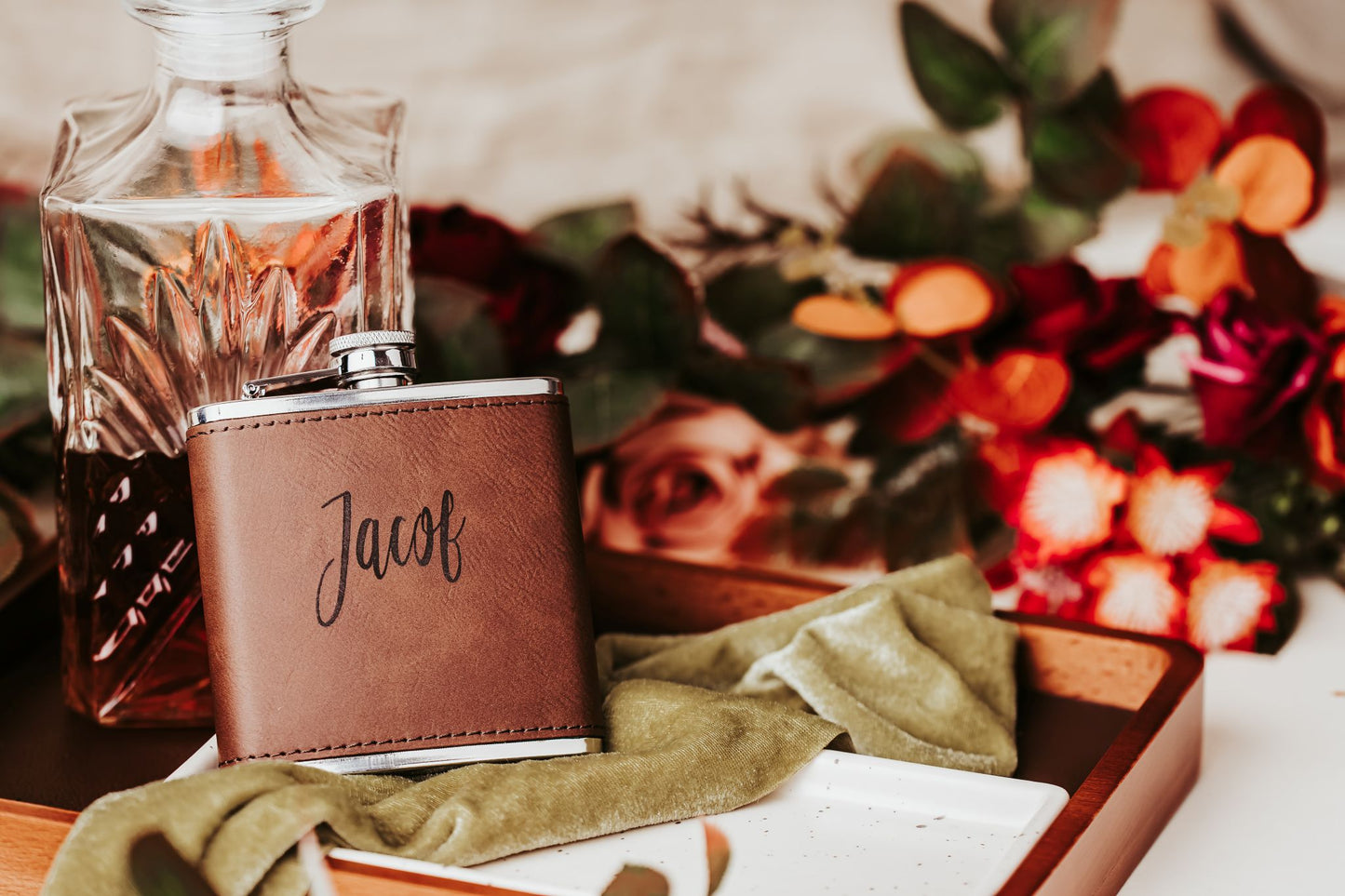 The Custom Engraved Flask by Mirabilia Boutique—a personalized leather gift for him—features the name "Jacob" and sits next to a glass decanter and green cloth, accented by colorful flowers; a perfect groomsmen gift.