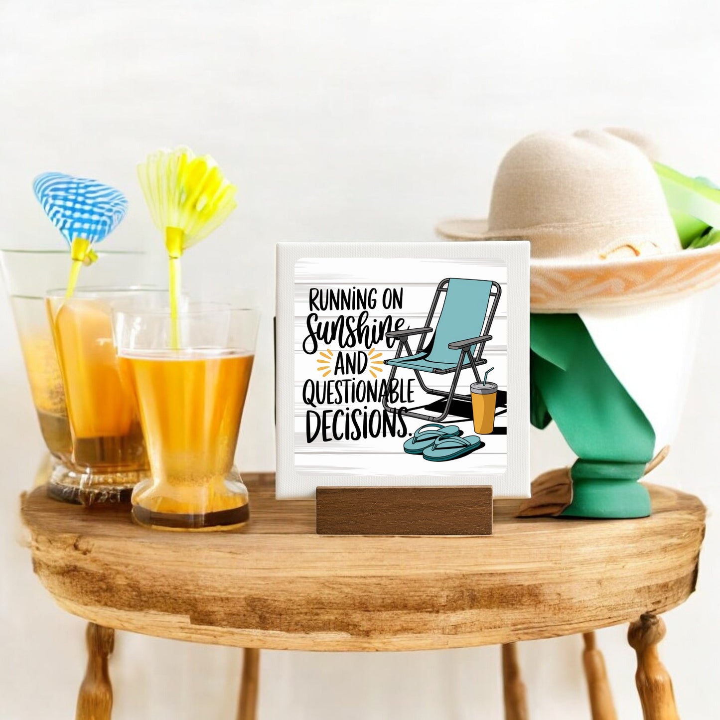 A small wooden table displays two tropical drinks, a sunhat with a green ribbon, and Mirabilia Boutique’s 4x4 “Running on Sunshine” mini canvas summer sign featuring a beach chair and flip-flops.