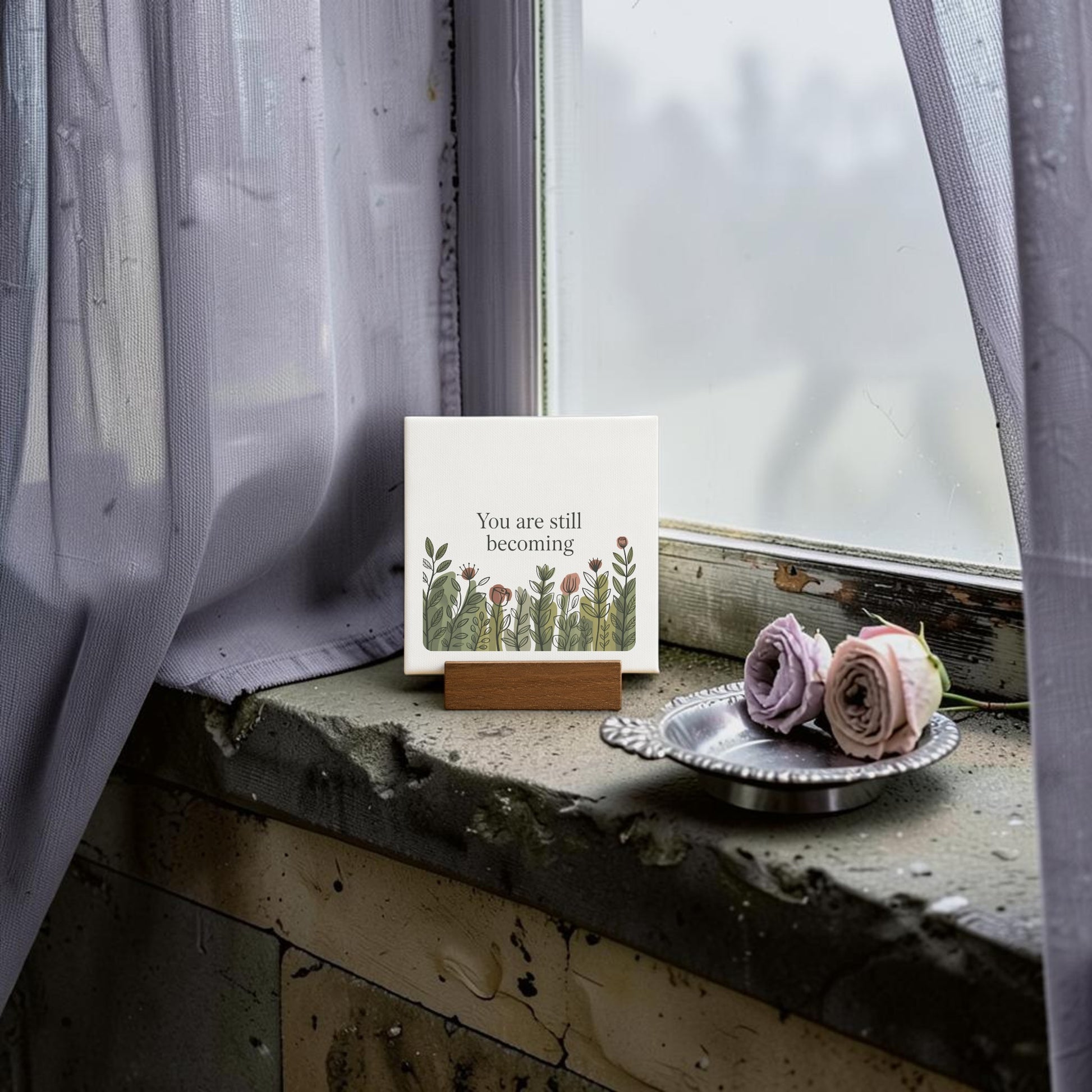 The Mirabilia Boutique "You Are Still Becoming" Mini Canvas Sign featuring floral illustrations sits on a windowsill beside mauve roses in a silver tray, framed by misty glass and sheer purple curtains for a soft botanical touch.