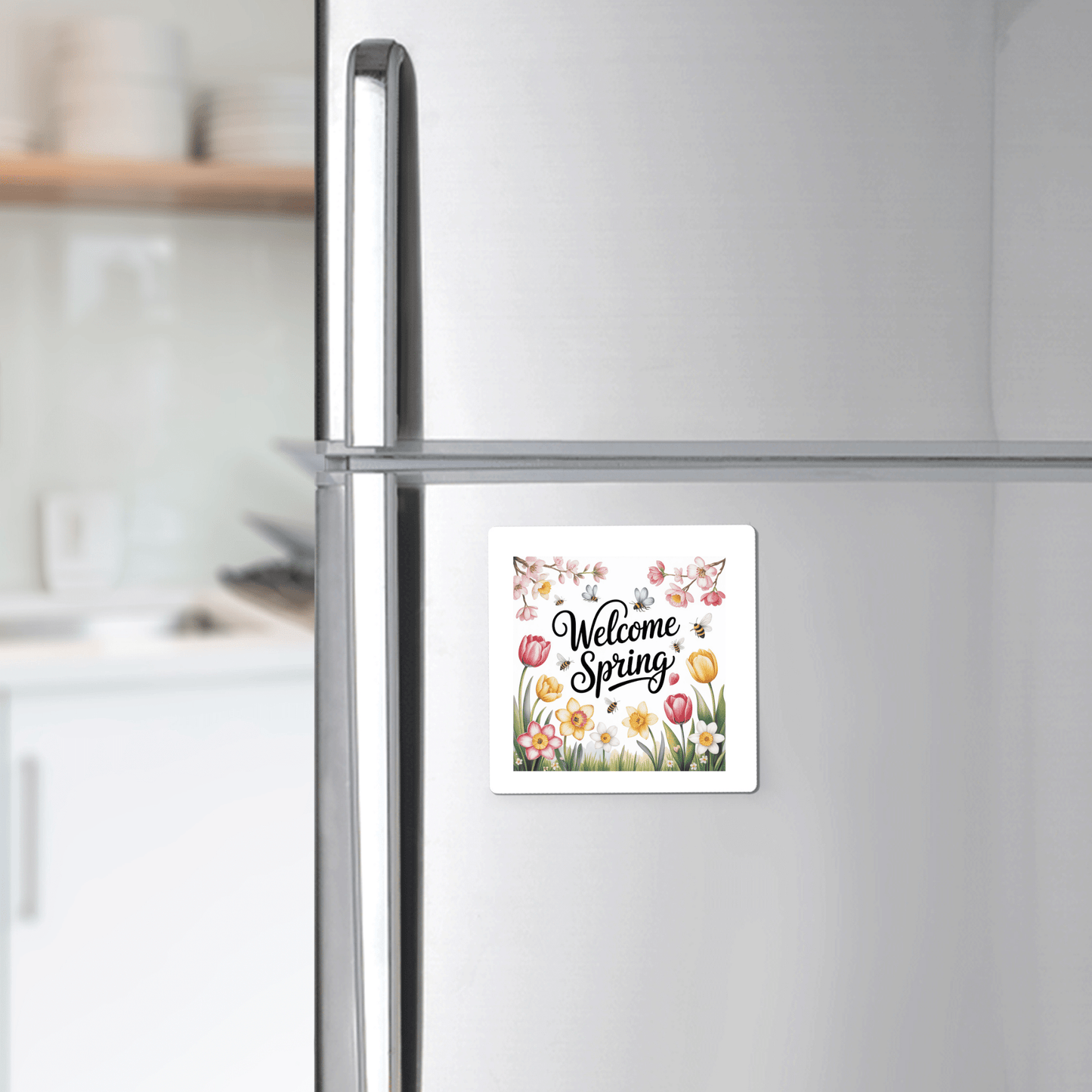 A stainless steel fridge displays the Mirabilia Boutique Welcome Spring Magnet – Floral Garden and Bees, featuring flowers and bees. In the background, kitchen shelves with dishes complete the cheerful spring atmosphere.