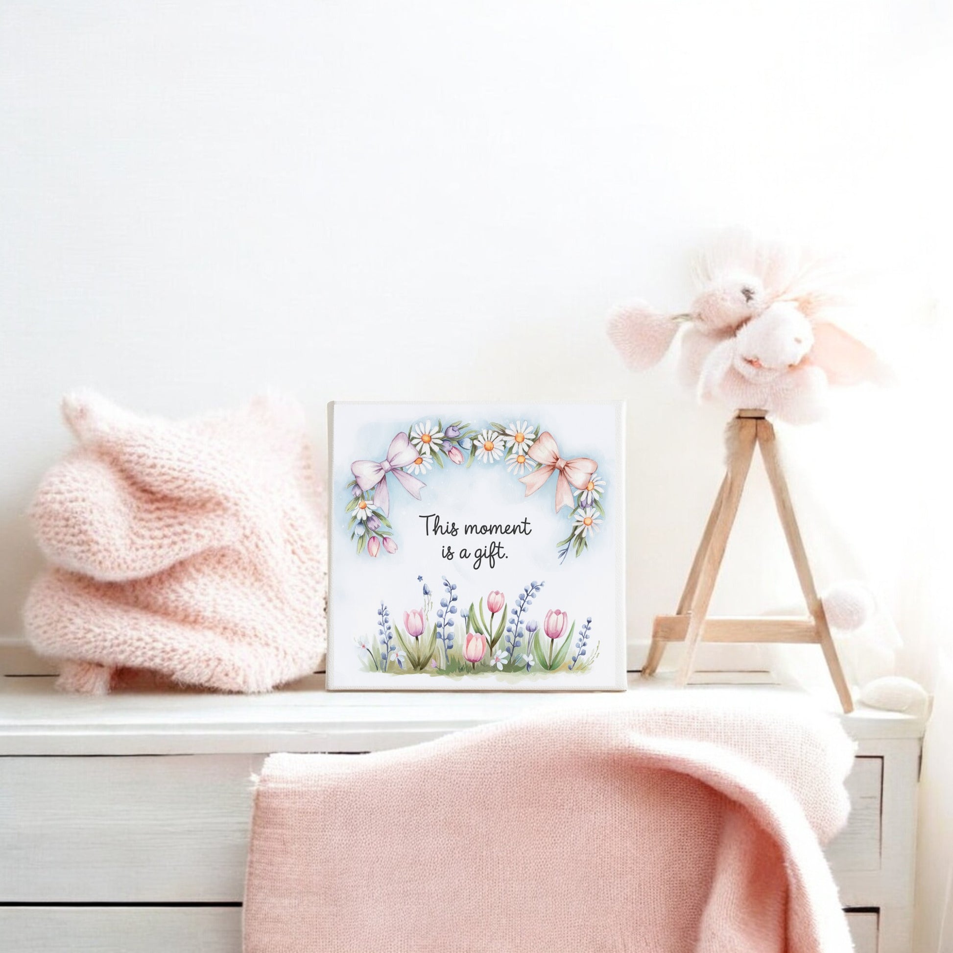 A pastel-colored nursery scene features a soft pink blanket, a knitted item, and a plush pig on a wooden stand. The "This Moment Is a Gift Canvas – 8x8 Spring Floral Art" by Mirabilia Boutique with floral accents hangs on the wall.