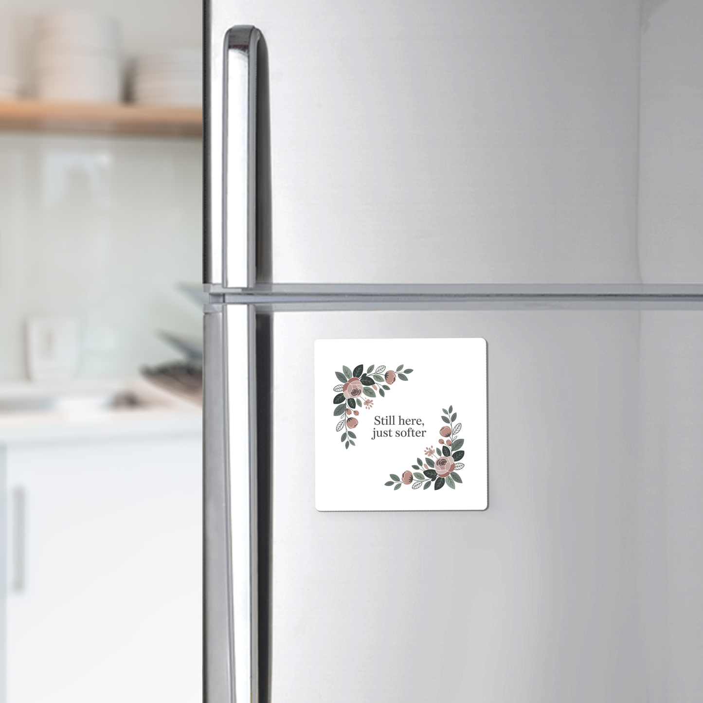 A silver fridge displays the "Still Here Just Softer Floral Magnet" by Mirabilia Boutique, featuring a botanical design and gentle message—an ideal encouragement gift for any softly blurred kitchen scene.